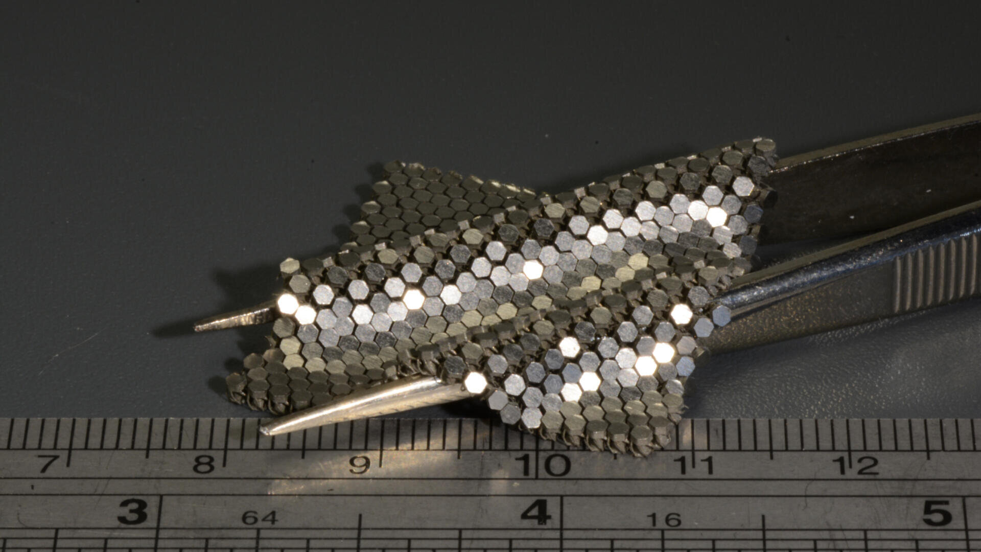 Eine Pinzette hält zwei kleine, sechseckige Metallgitterstrukturen in Flugzeugform. Ein Lineal darunter zeigt, dass die Strukturen etwa drei Zentimeter lang sind. Der Hintergrund ist eine schlichte, dunkle Fläche.