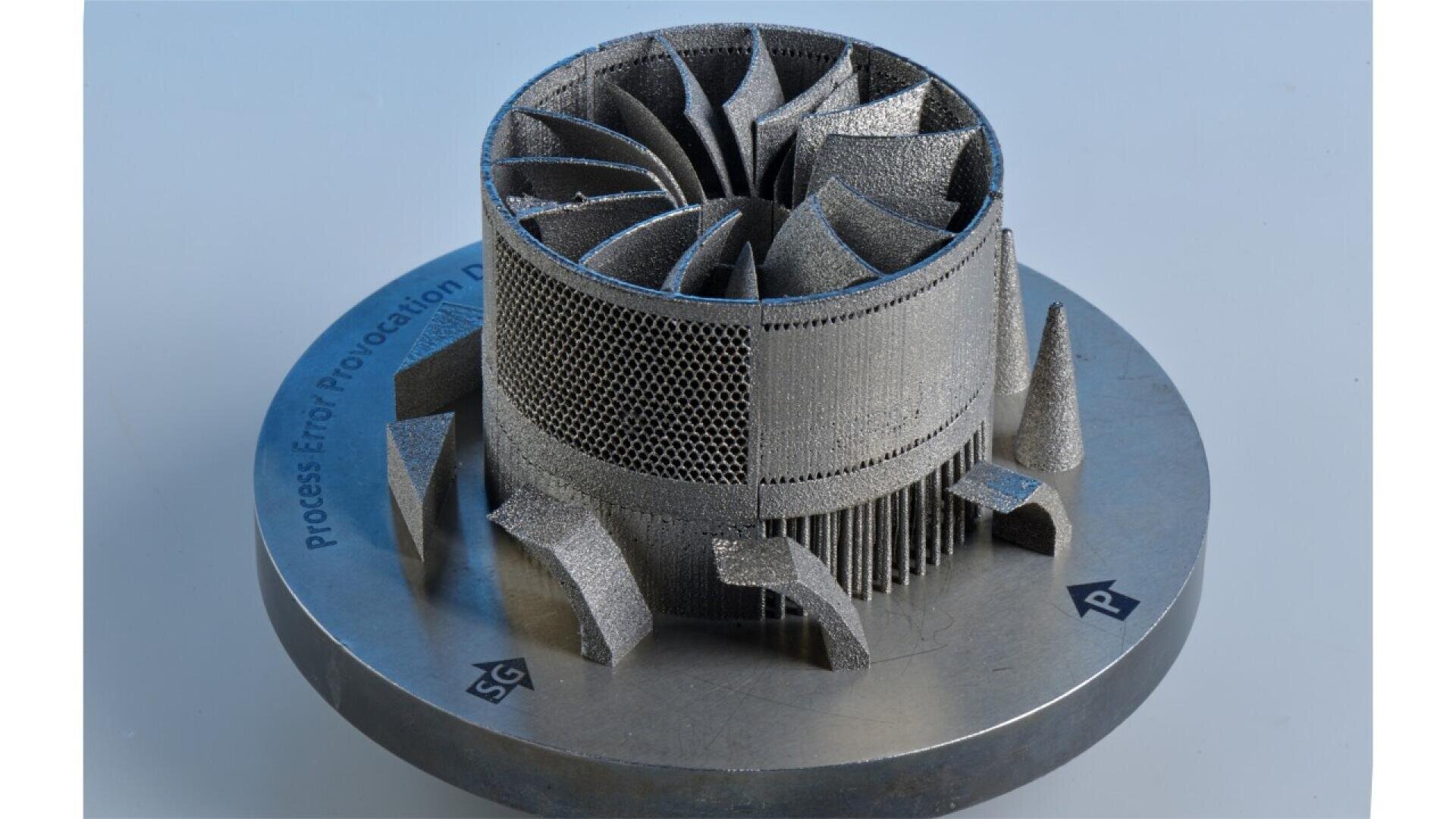 A metallic 3D-printed turbine component with curved blades and detailed mesh structures stands on a round metal base, displayed against a light blue background.