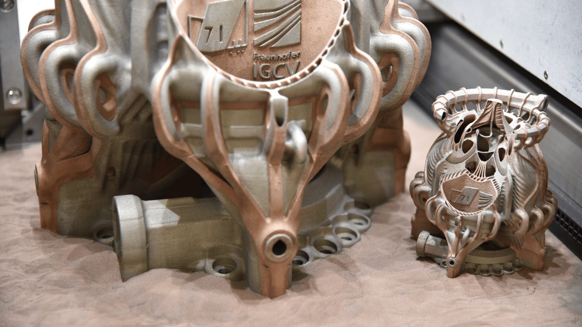 Zwei verschlungene 3D-gedruckte Metallobjekte mit komplexen, organischen Formen und geprägten Logos liegen auf einem Sandbett in einer industriellen Umgebung. Die Objekte weisen eine Mischung aus metallischen Silber- und Kupfertönen auf.