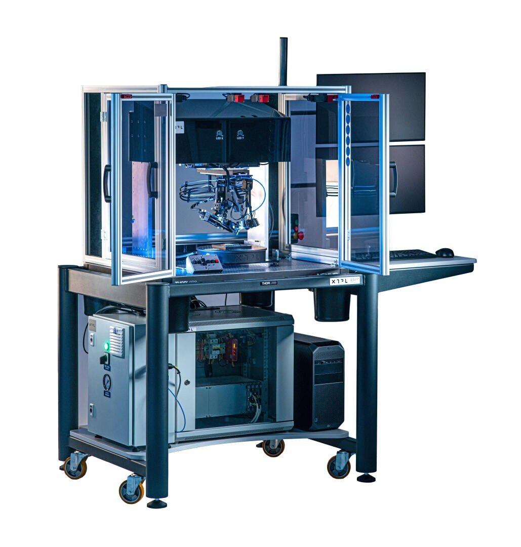 Eine hochtechnologische automatisierte Labormaschine mit einem Roboterarm in einem Schutzgehäuse, mehreren Monitoren und Computerhardware auf einem Tisch mit Rollen, die für wissenschaftliche Experimente oder die Fertigung verwendet wird.