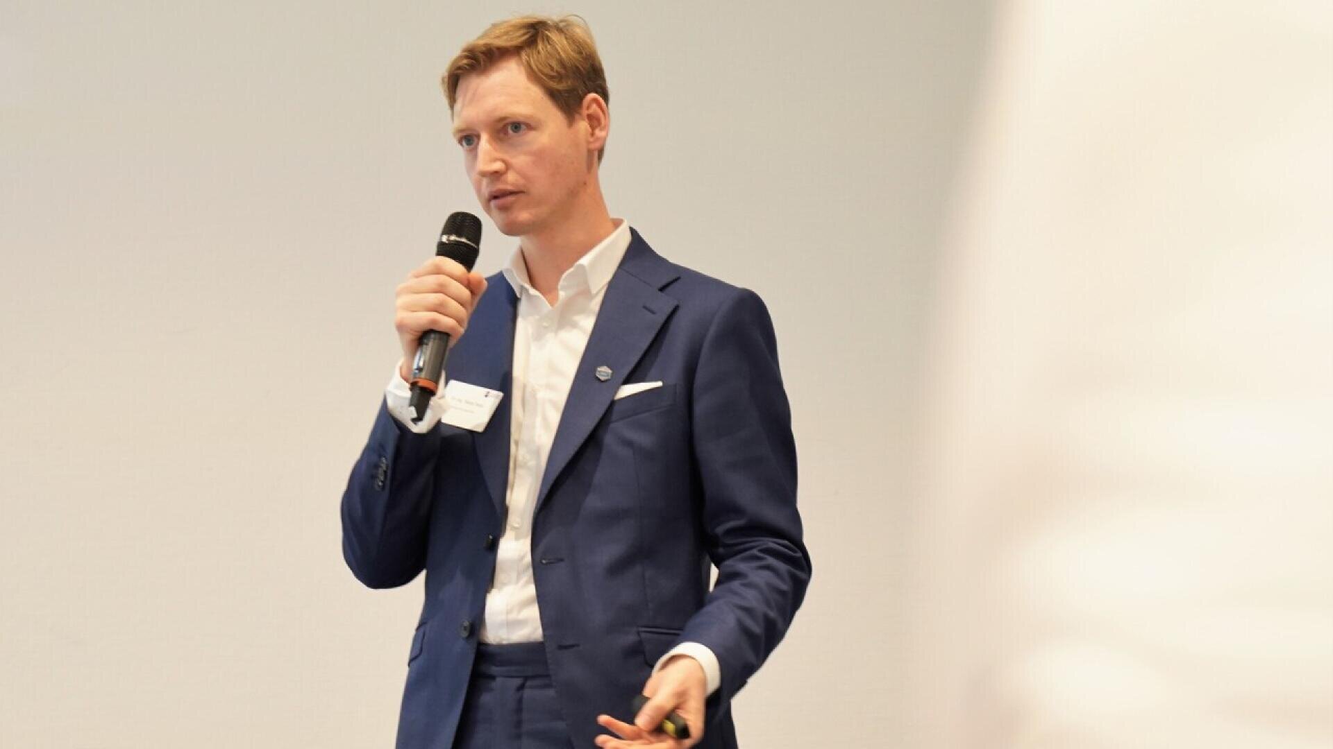 A man in a blue suit holds a microphone and speaks, standing against a plain light background. He appears to be giving a presentation, with a name tag visible on his jacket.