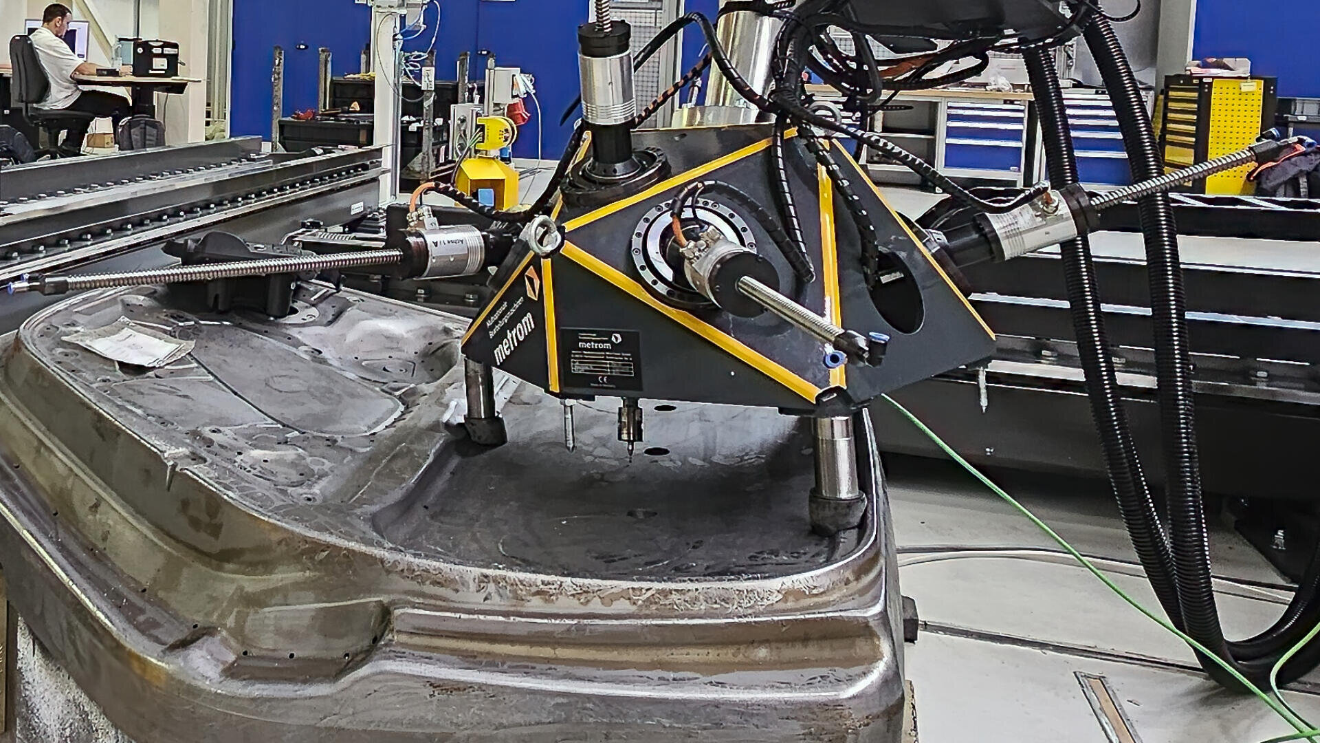 A large coordinate measuring machine with a probe inspects a metal mold in an industrial workshop, surrounded by equipment and tool cabinets.