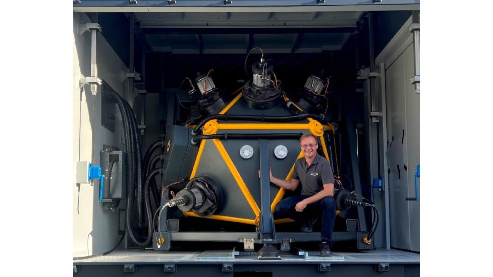 A person squats next to a large industrial machine with yellow and black components, housed inside a container. The machine features multiple hoses and mechanical parts, and the person is smiling at the camera.