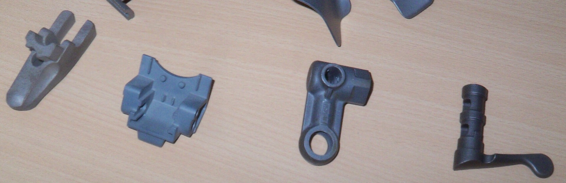 Vier verschiedene mechanische Metallteile liegen in einer Reihe auf einer hellen Holzoberfläche. Jedes Teil hat eine eigene Form und scheint aus dunkelgrauem Metall zu bestehen.