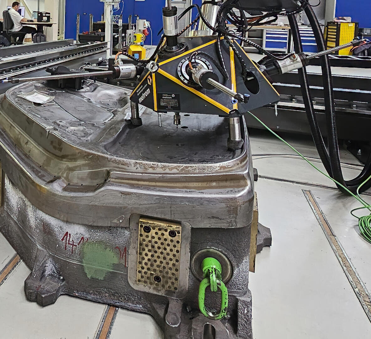 Eine große Industriemaschine mit montiertem Koordinatenmessgerät prüft ein schweres Metallteil in einer mit verschiedenen Werkzeugen und Geräten ausgestatteten Werkstatt.