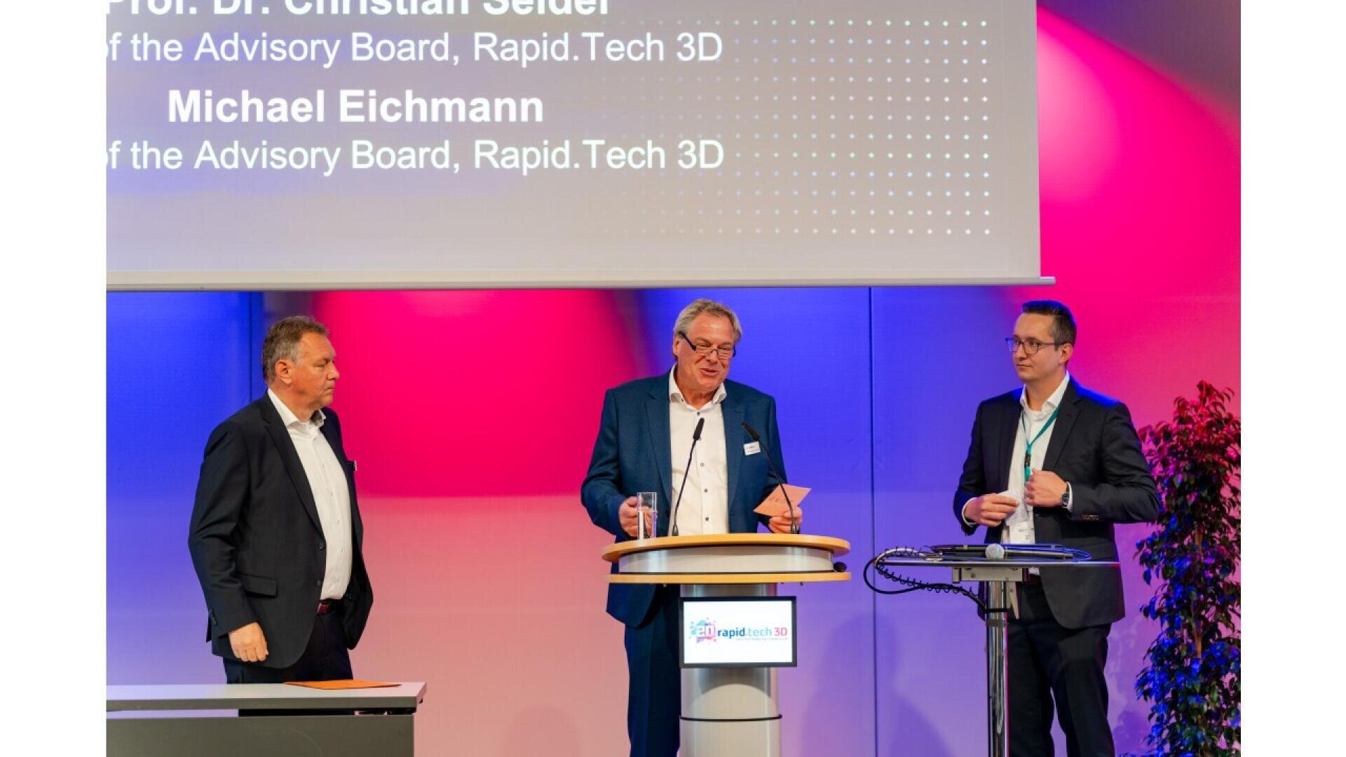 Three men in business attire stand on a stage having a discussion. One man speaks at a podium, while the other two listen. A large screen behind them displays names and titles related to Rapid.Tech 3D.