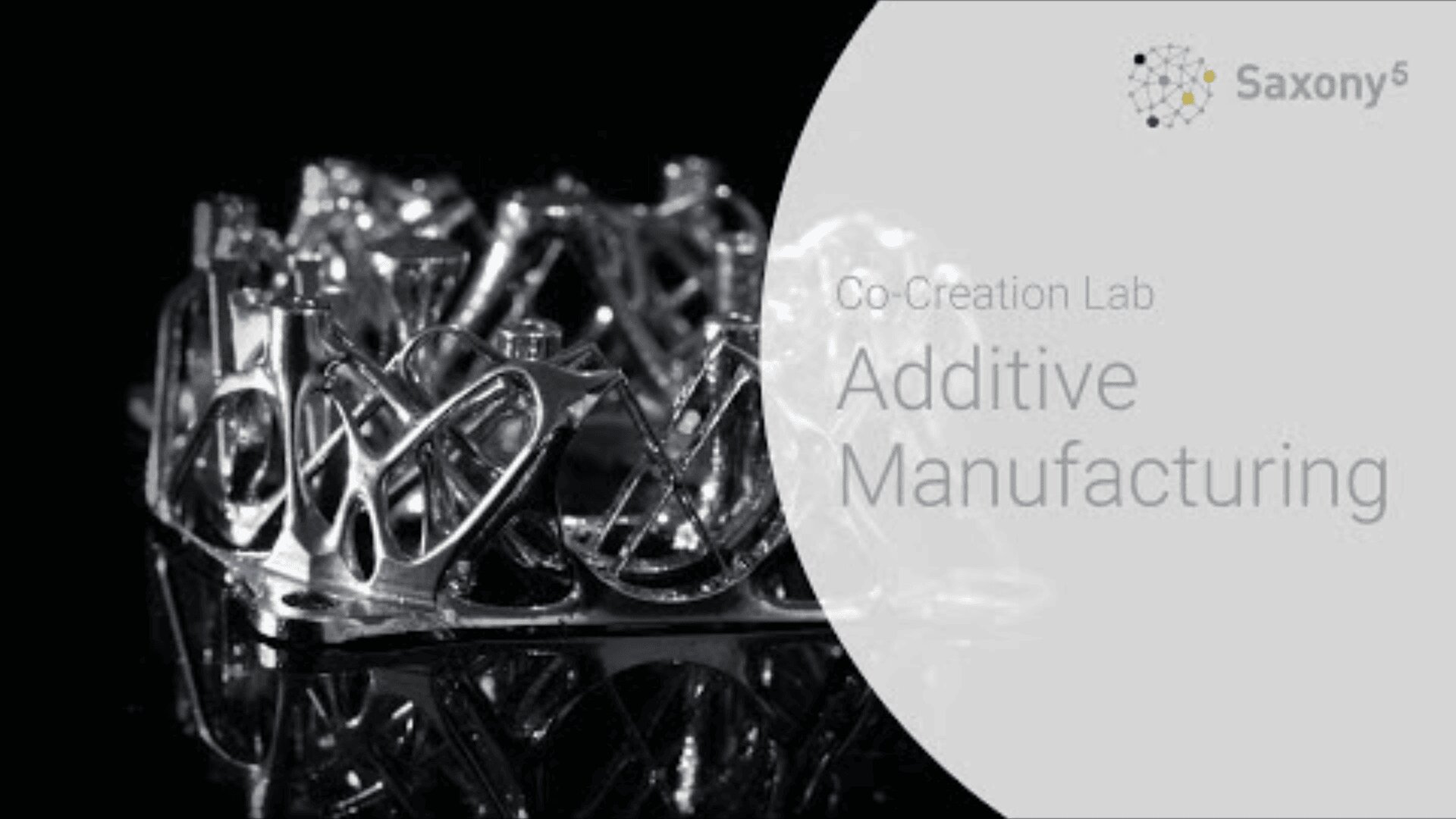 A metallic, lattice-like 3D printed object is displayed on a black surface. Overlaid text reads: Co-Creation Lab Additive Manufacturing with the Saxony^5 logo in the upper right corner.