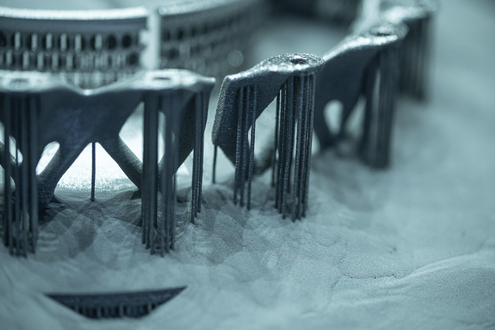 Nahaufnahme eines metallischen 3D-gedruckten Objekts mit komplizierten Gitterstrukturen, Stützsäulen und pulverförmigen Rückständen rund um die Basis, was auf eine Industrie- oder Fertigungsumgebung hindeutet.