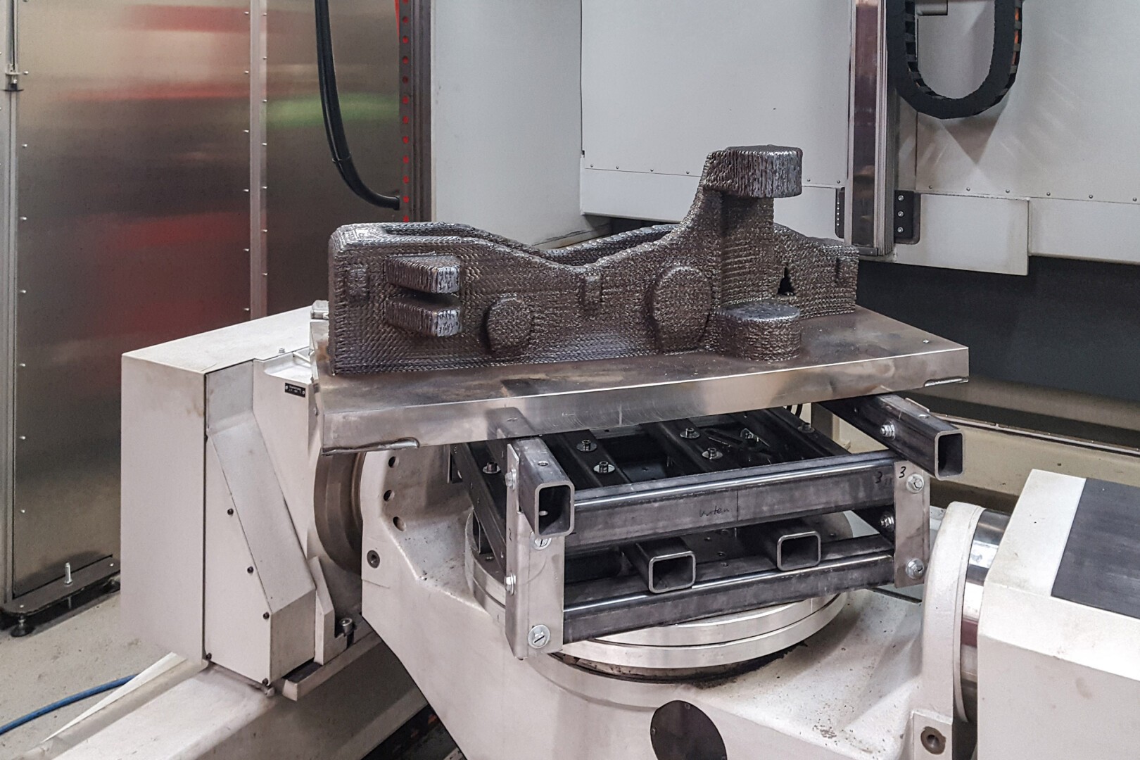 Ein großes, im 3D-Druckverfahren hergestelltes Industrieteil aus Metall befindet sich auf einer rotierenden Plattform in einer Fertigungsmaschine, umgeben von mechanischen Komponenten und metallischen Oberflächen.