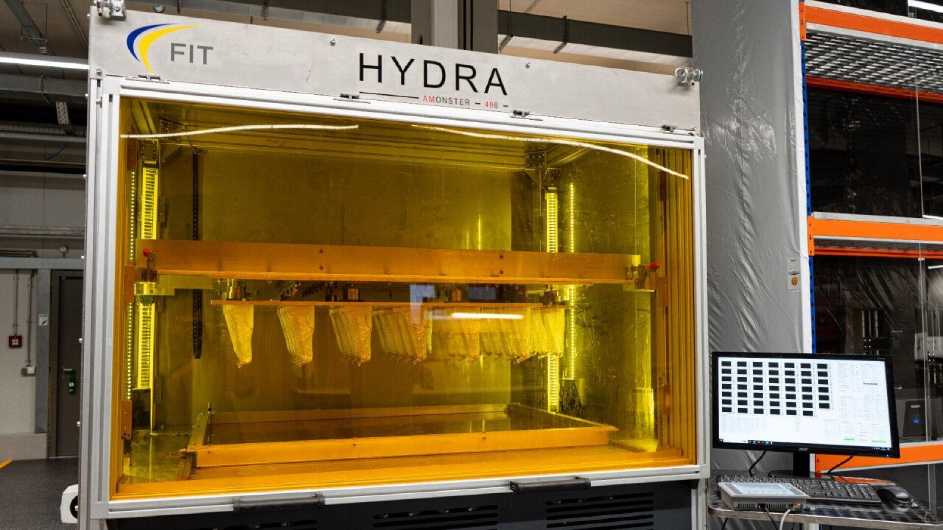 A large industrial 3D printer labeled HYDRA with a yellow-tinted enclosure is in operation, producing multiple parts. A computer monitor with a grid display stands beside the machine in a modern workspace.