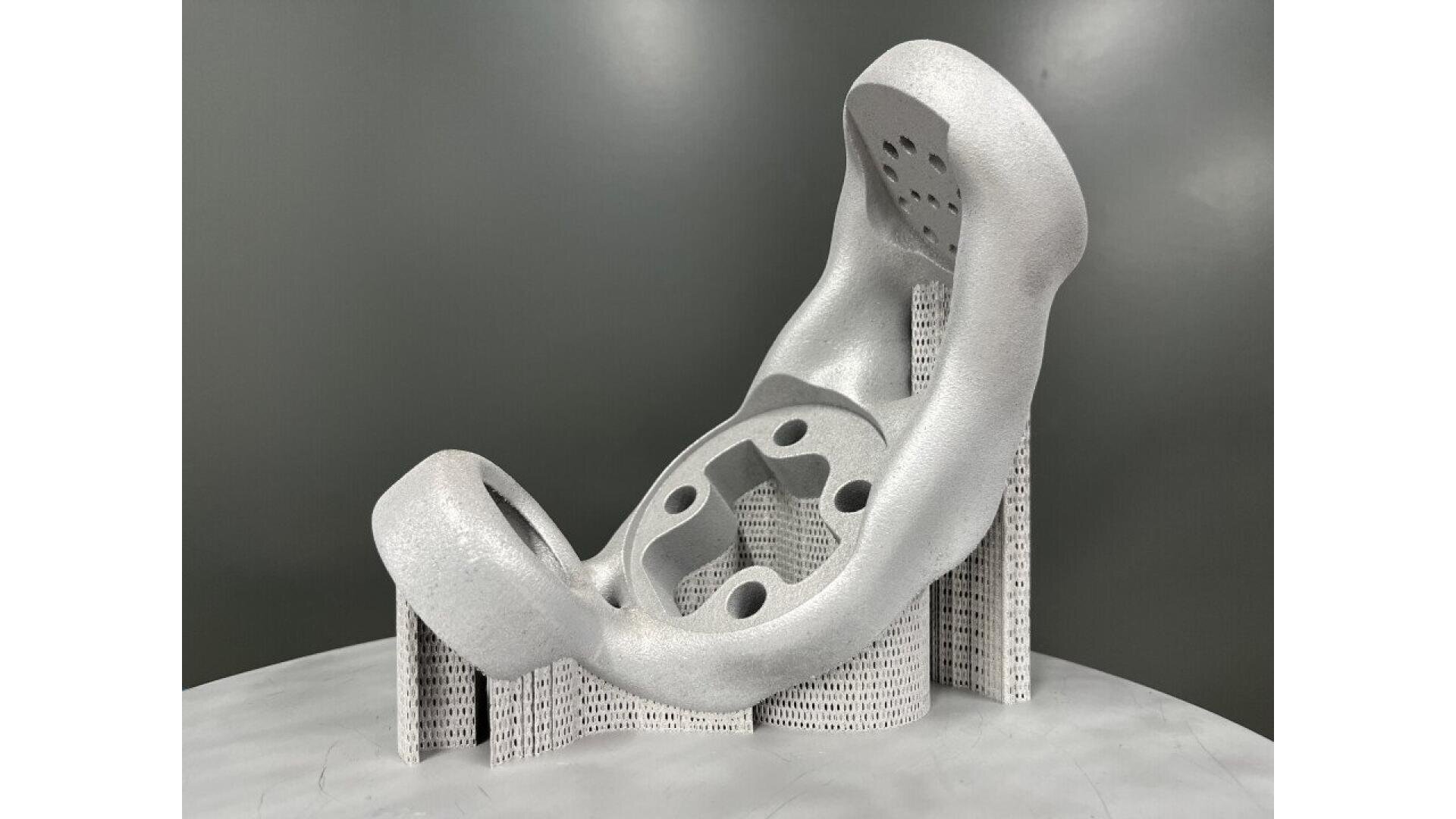 A complex, metallic 3D-printed part with curved, hollow sections and multiple holes, supported by a lattice-like structure, displayed against a plain gray background.