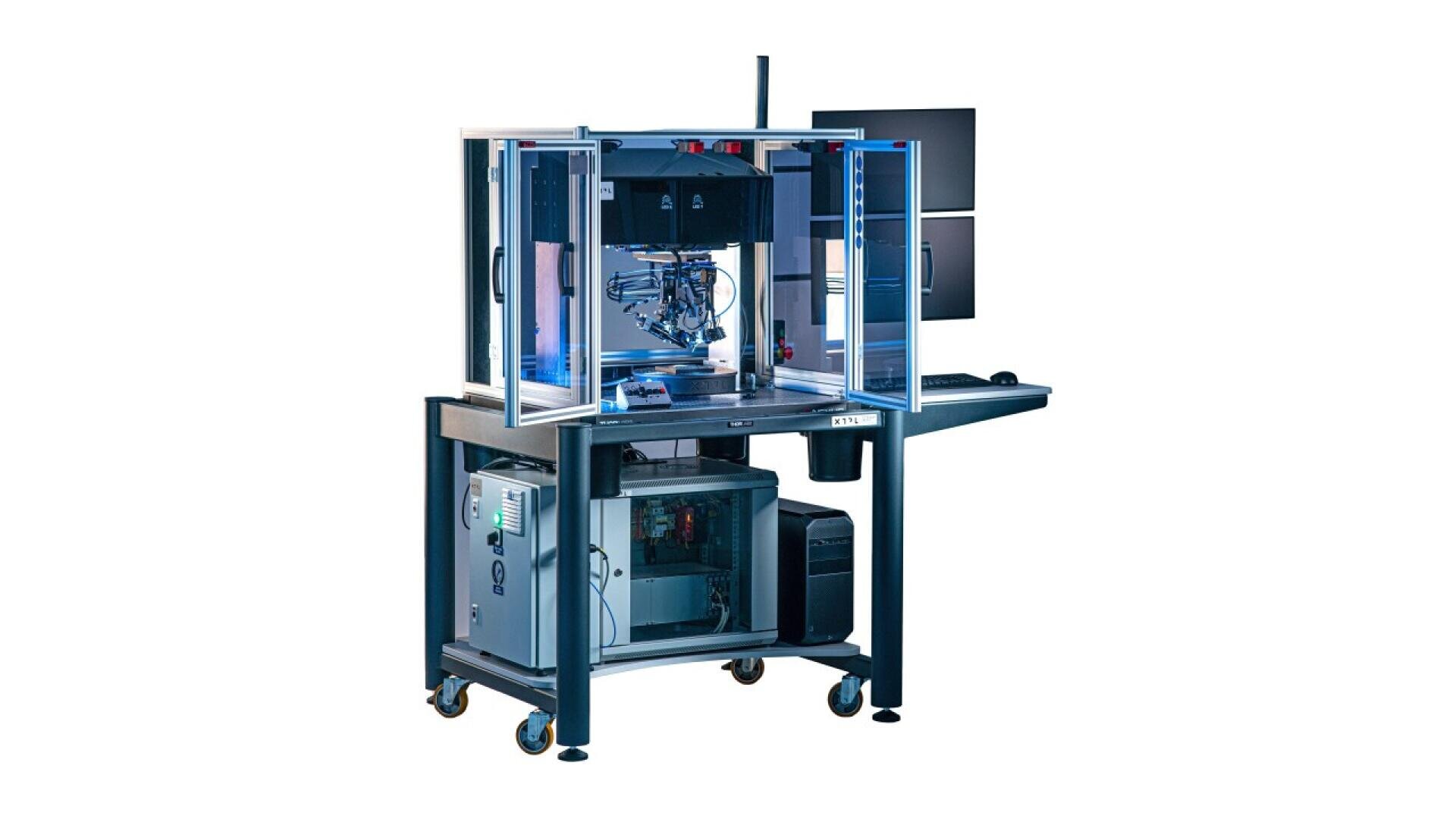 A computer-controlled laboratory device with a transparent enclosure sits on a wheeled table, featuring visible mechanical components, wires, and monitors in the background.