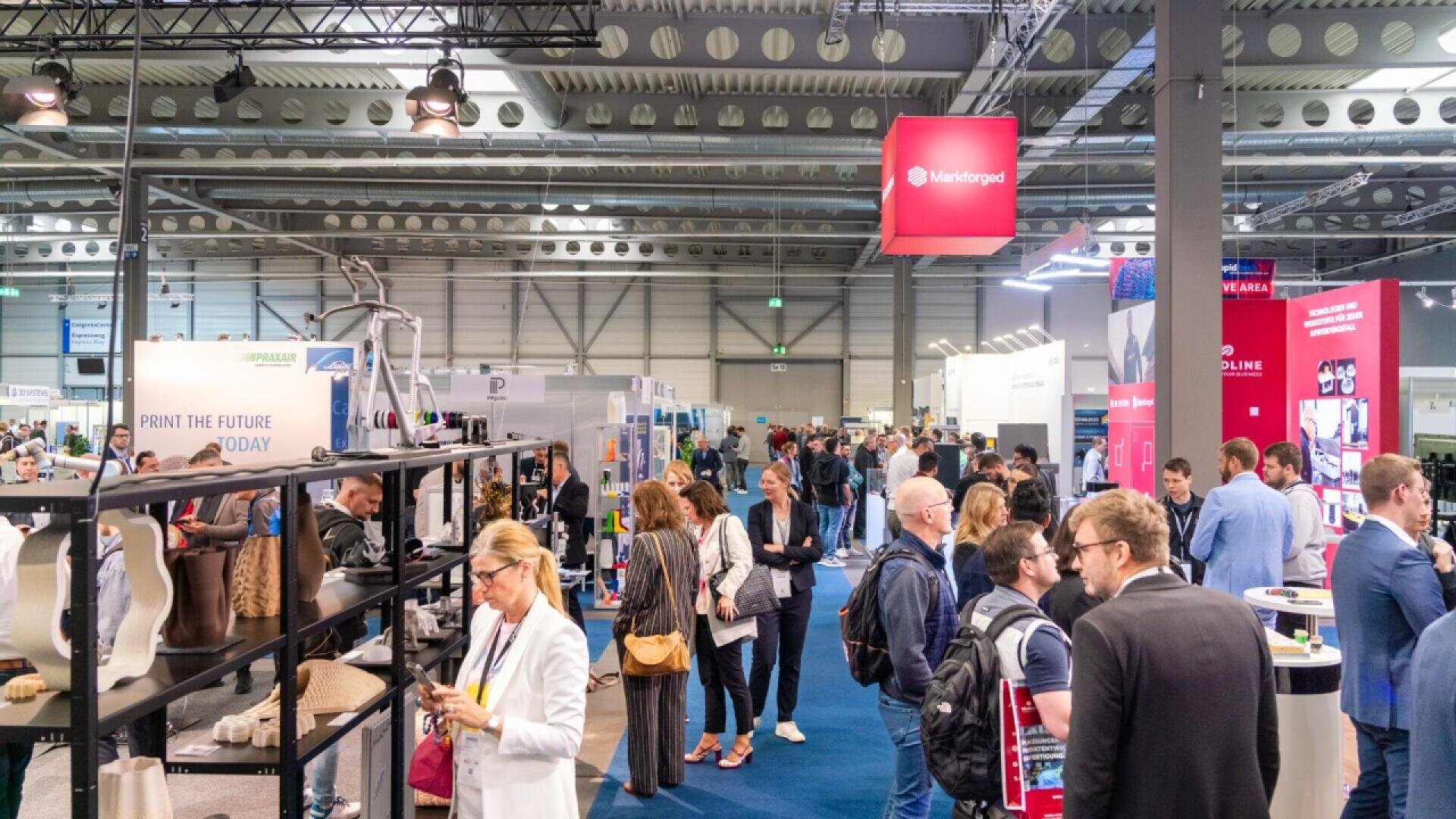A busy trade show floor filled with people networking and visiting various exhibition booths under bright industrial lighting in a spacious convention hall.