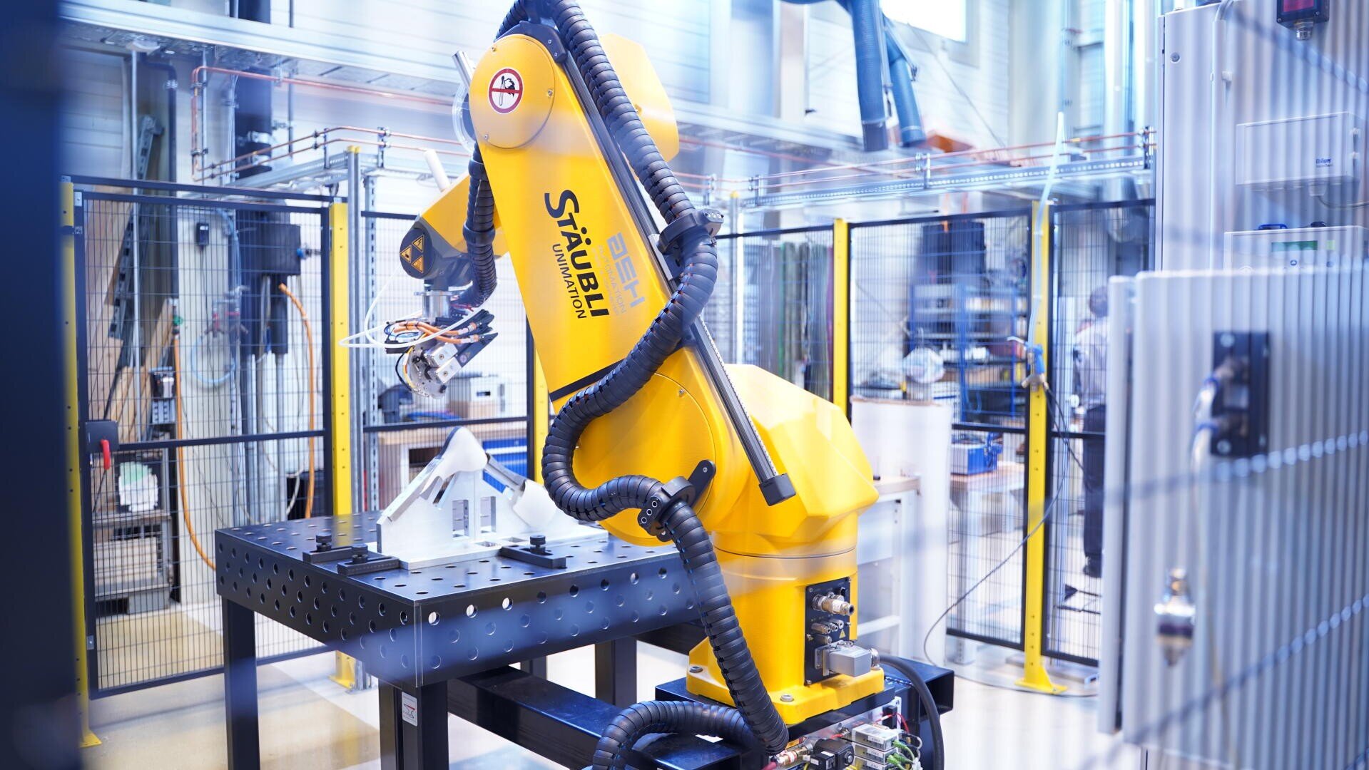 Ein großer gelber Industrieroboterarm arbeitet in einer modernen Fabrik an einem Metallarbeitsplatz mit sichtbarer technischer Ausrüstung und Verkabelung.