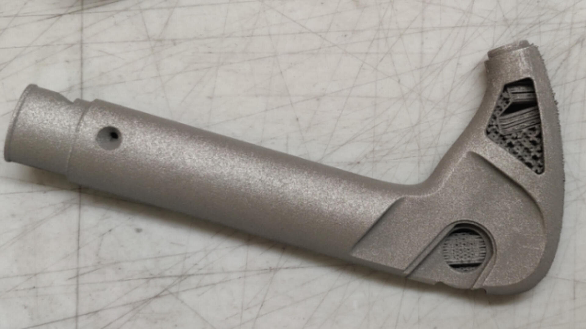 Ein metallisches, L-förmiges 3D-gedrucktes Teil mit strukturierter Oberfläche und geometrischen Ausschnitten, das auf einer zerkratzten weißen Oberfläche ruht.