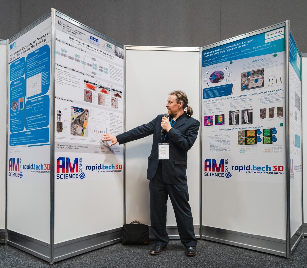 A man in a suit holds a microphone in his hand and points to a scientific poster at a conference. He is standing between two display boards with technical diagrams and images, under banners reading rapid.tech 3D and AM Science.