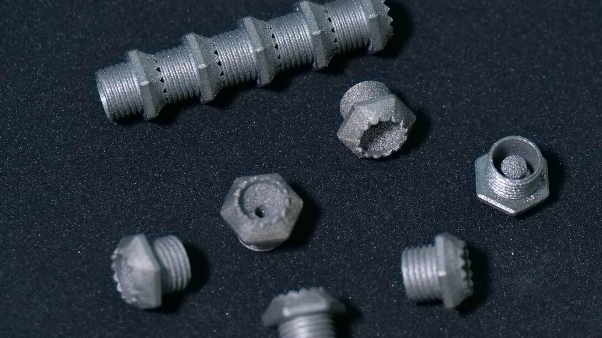 Mehrere kleine, graue, sechseckige Metallschrauben und -muttern sind auf einer schwarzen Oberfläche verstreut, mit einem längeren Gewindestück oben im Bild.