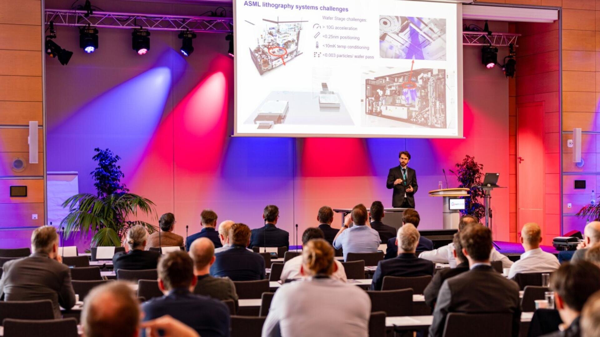 A speaker presents a slideshow about ASML lithography system challenges to an audience in a conference room with colorful lighting and seated attendees facing the stage.