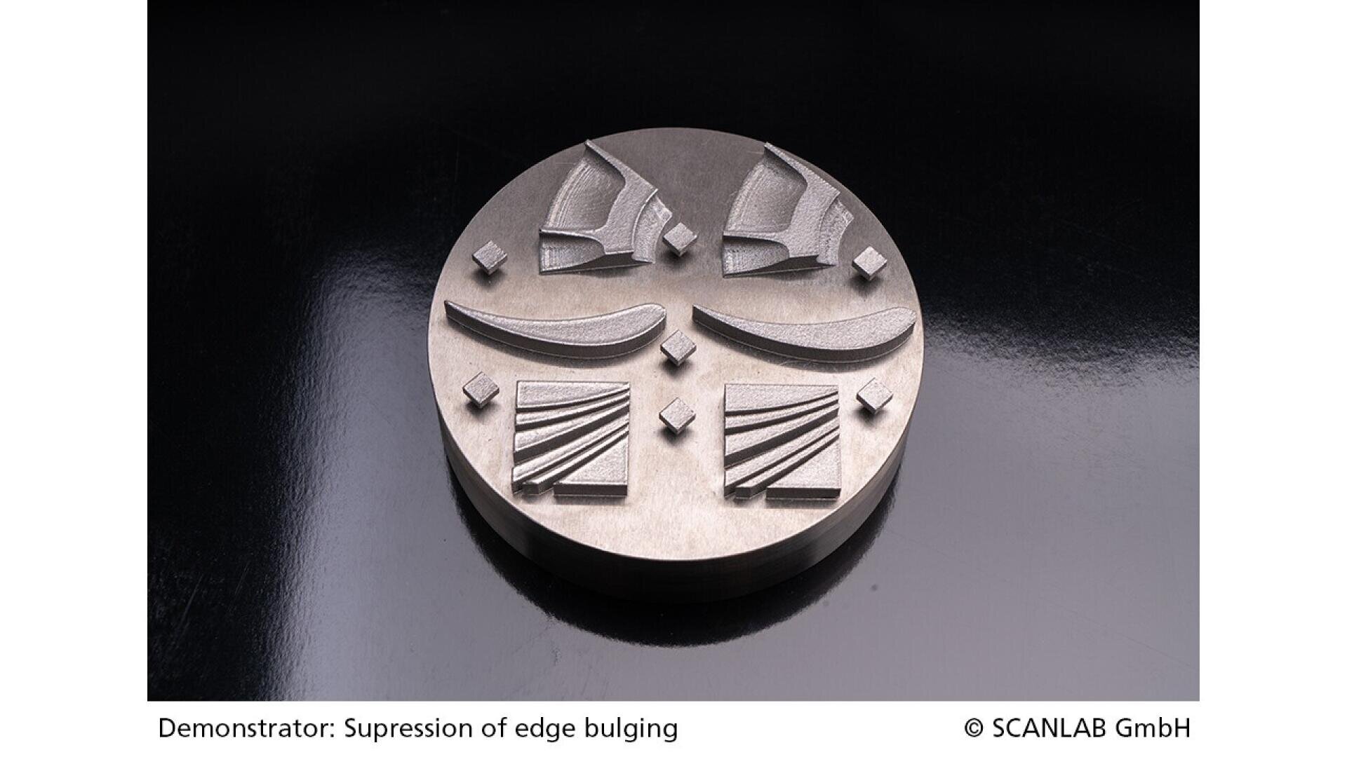 A round metallic object displays four raised geometric patterns and shapes, demonstrating the suppression of edge bulging on its smooth surface. The object is placed on a glossy black background.