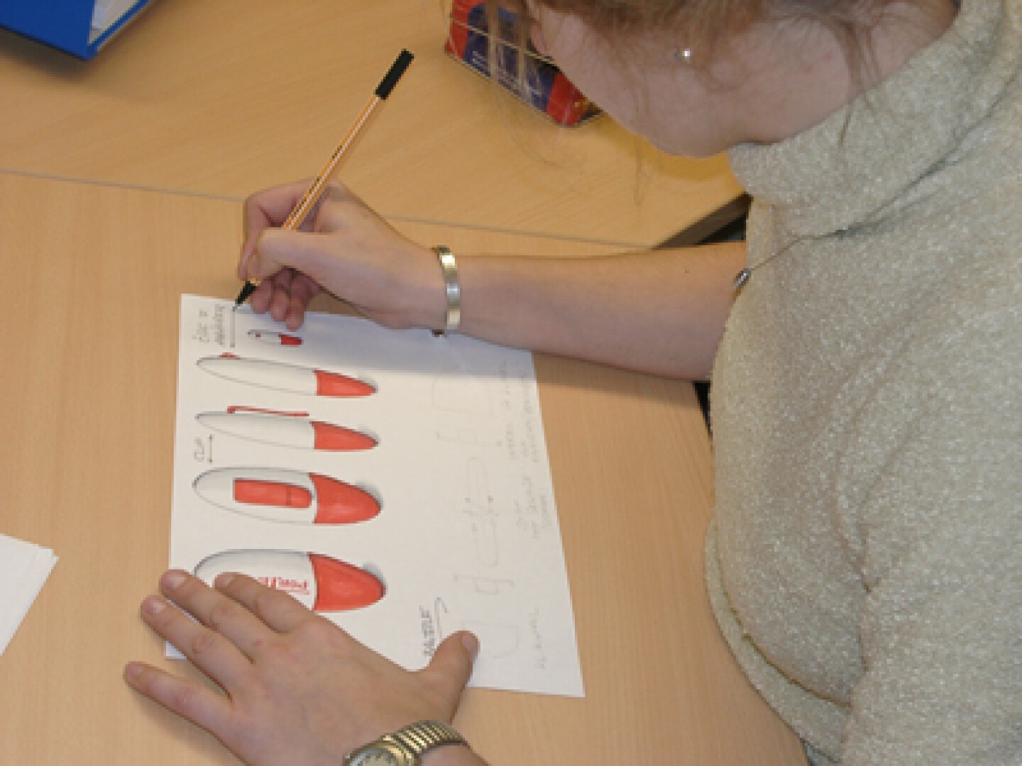 Eine Person skizziert und malt rote und weiße Designs ergonomischer Computermäuse auf einem Blatt Papier, während sie an einem Schreibtisch sitzt.