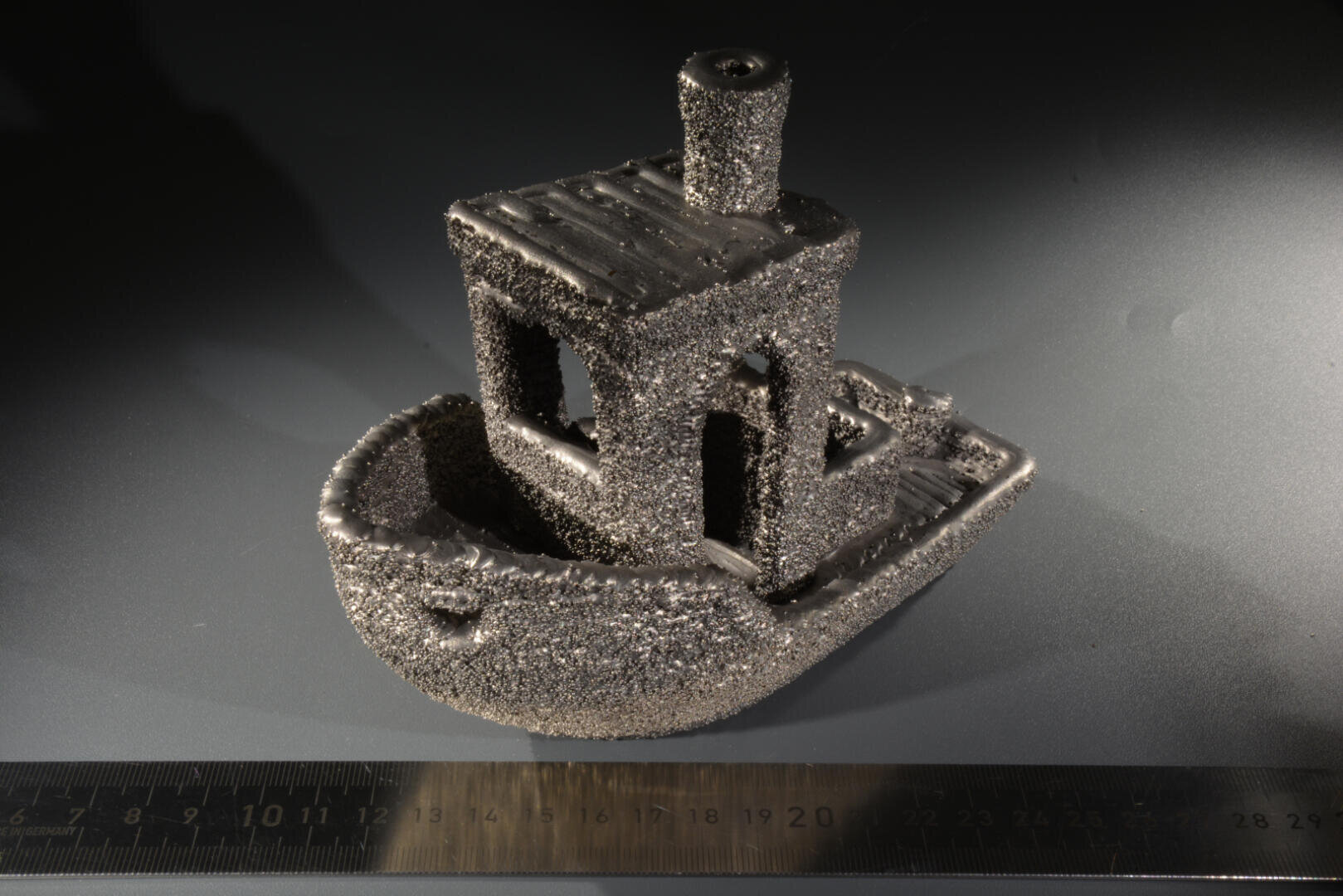 Ein kleines, grob strukturiertes 3D-gedrucktes Bootsmodell liegt auf einer dunklen Oberfläche neben einem Lineal und zeigt seine Größe. Das Boot wirkt metallisch und körnig, mit auffälligen Oberflächenfehlern.