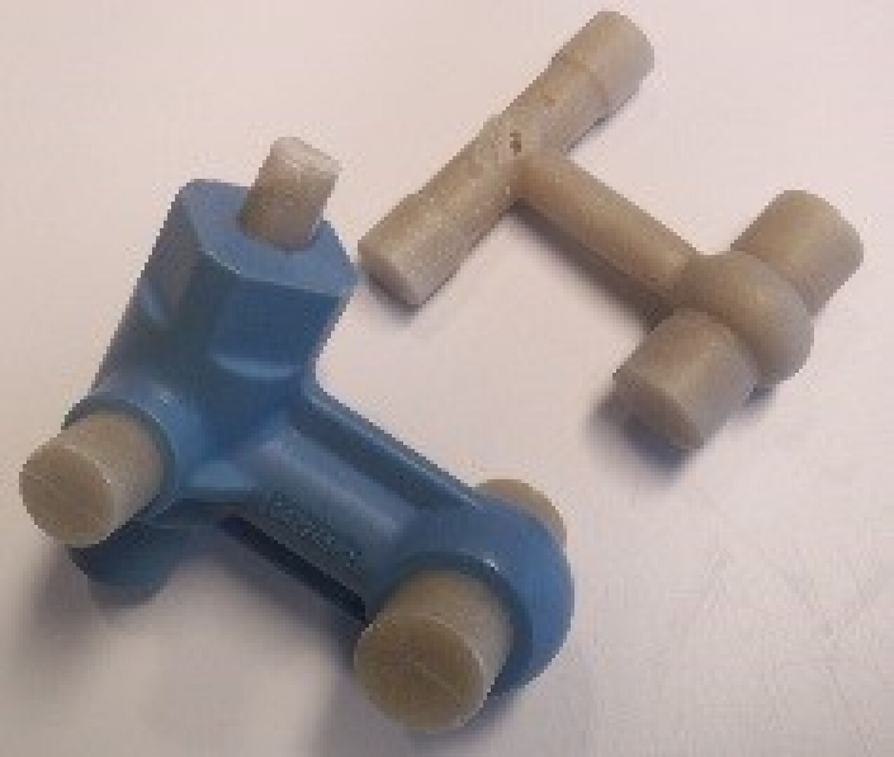 Zwei mechanische Kunststoffteile, eines blau und eines beige, jeweils mit zylindrischen Verlängerungen und T-förmigem Design, auf einer weißen Oberfläche platziert.