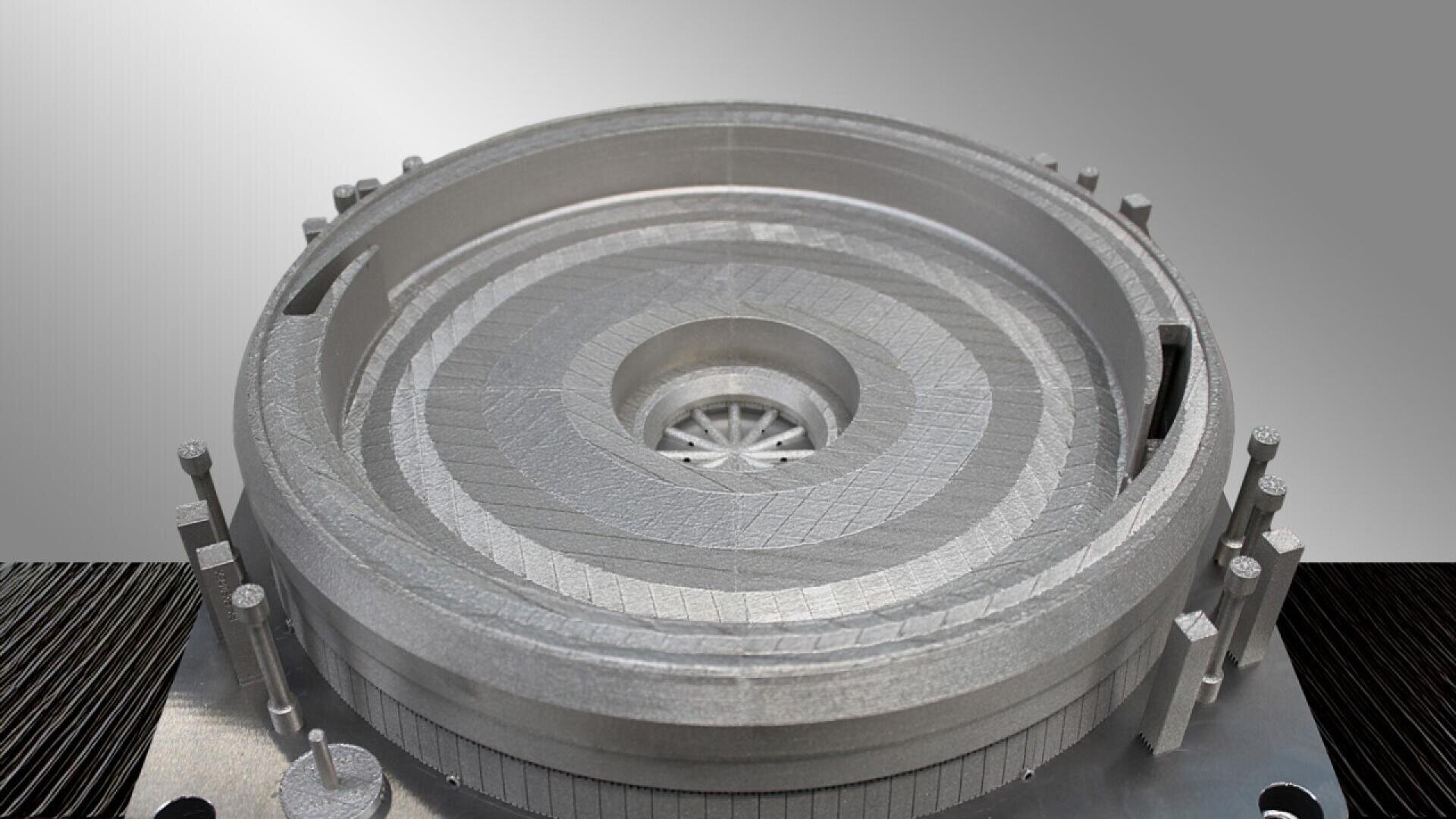 A close-up view of a metallic, circular, 3D-printed part with concentric layers and intricate internal structures, resting on a flat surface against a gradient gray background.