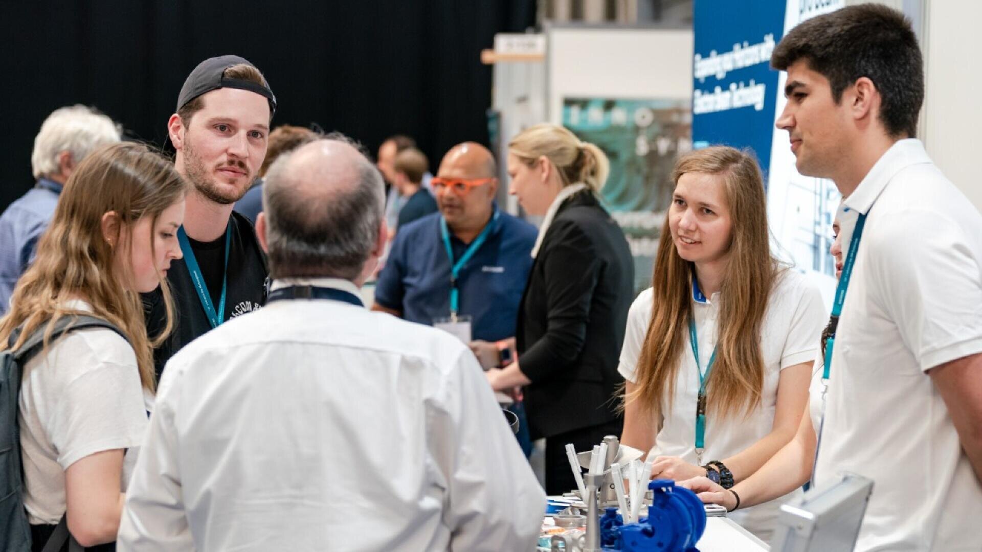 A group of young adults talks with an older man at an exhibition booth, with machines and brochures on the table. Other attendees interact in the background of the busy event.