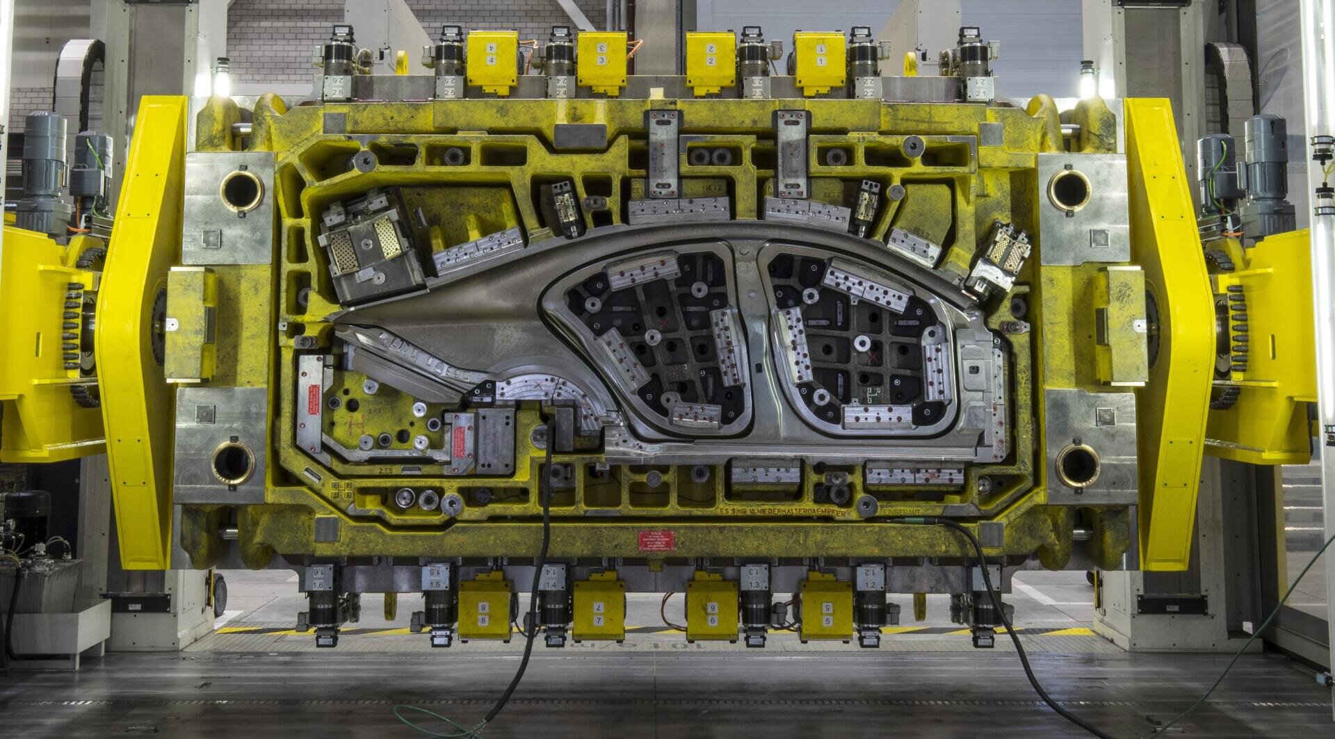 Eine große industrielle Stanzpresse mit gelbem Rahmen, die eine komplexe Metallform hält, die zum Formen von Karosserieteilen in einer Fabrik verwendet wird.