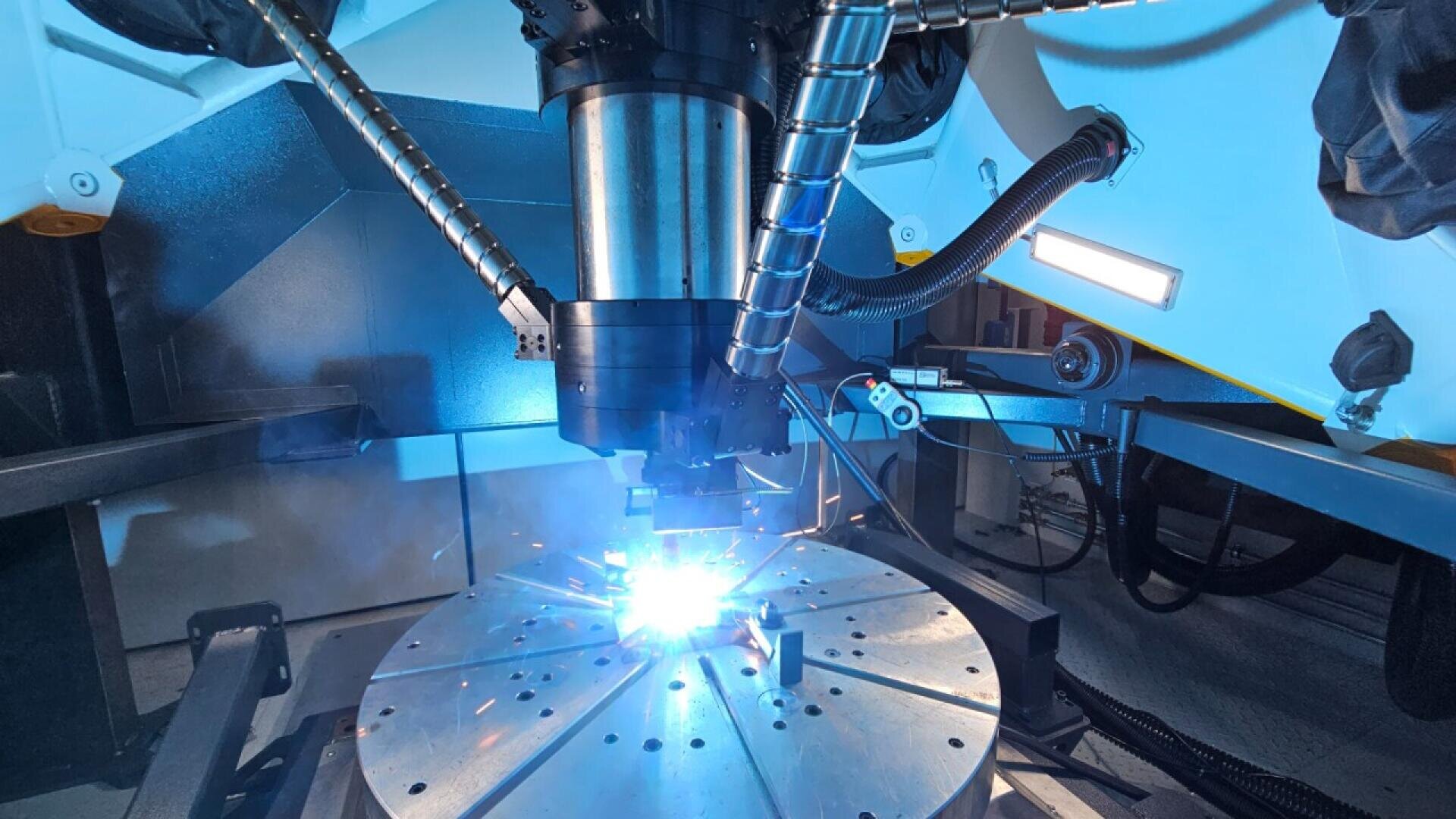 A close-up view of an industrial robotic machine welding a metal component, emitting bright blue sparks, inside a modern, high-tech manufacturing facility.