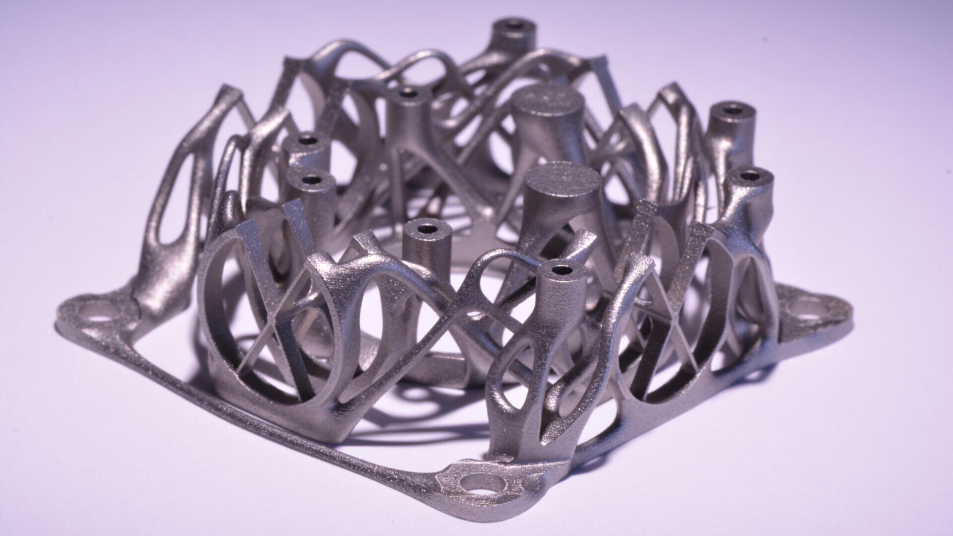 Eine Nahaufnahme eines metallischen 3D-gedruckten Objekts mit einer komplizierten, gitterartigen Struktur, die ineinander verwobene geometrische Formen und zylindrische Elemente aufweist und auf einer flachen Oberfläche mit einem violetten Farbton platziert ist.