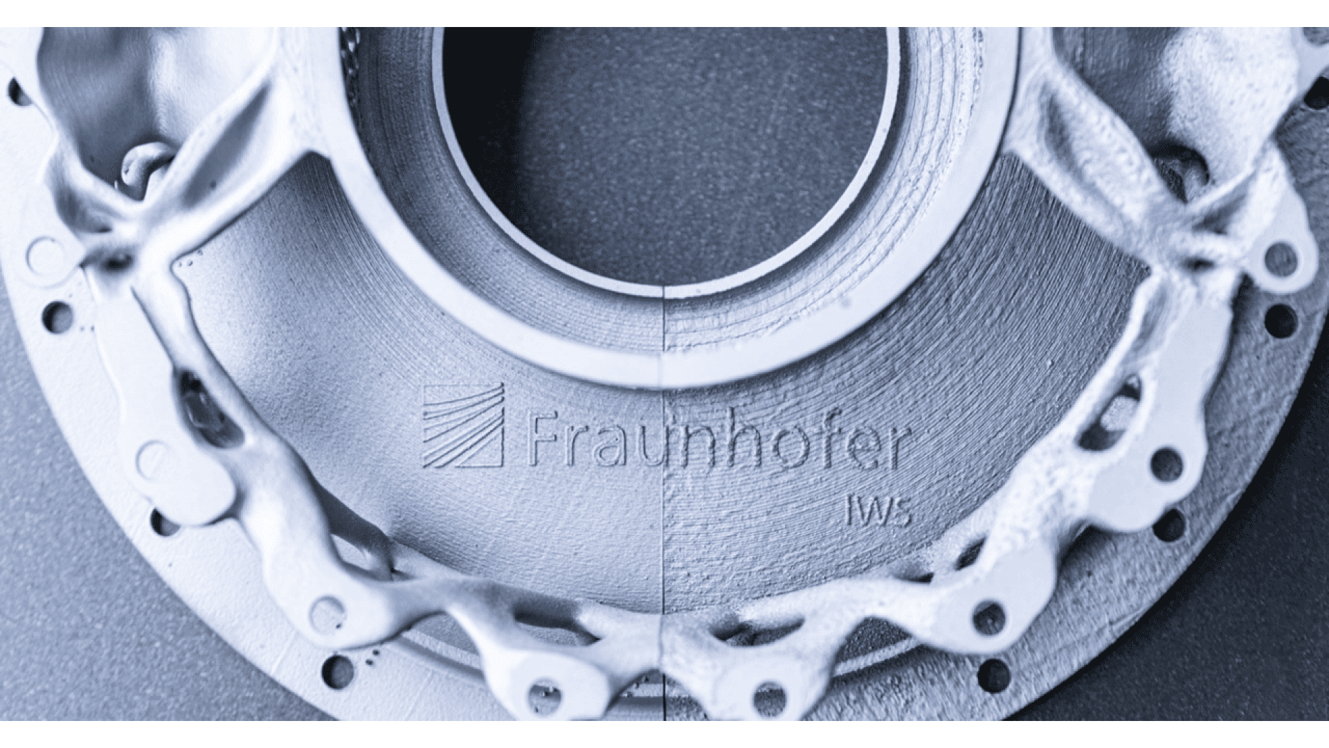 Close-up of a metallic, circular mechanical part with intricate design details, shown in two different surface finishes split vertically; “Fraunhofer IWS” is labeled across the center.