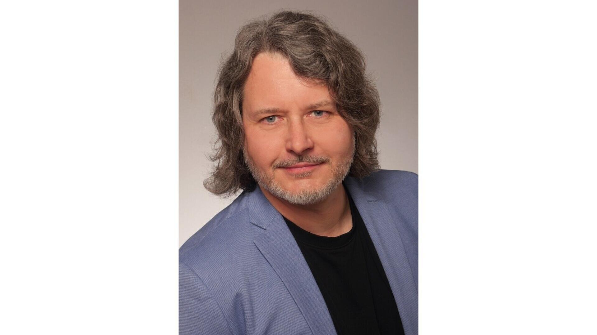A man with wavy, shoulder-length gray hair and a short beard is wearing a light blue blazer over a black shirt, posing against a plain, light background.