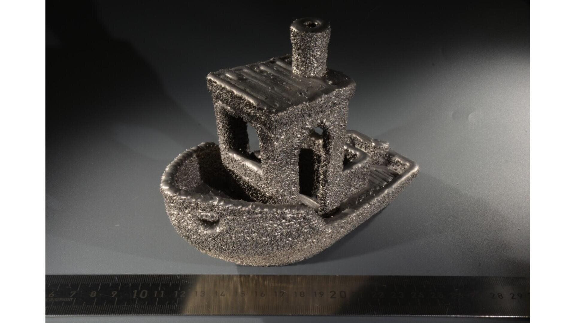 A metallic 3D-printed model of a small boat, covered in rough, sparkly texture, sits on a dark surface next to a black ruler showing centimeter measurements. The lighting creates noticeable shadows.
