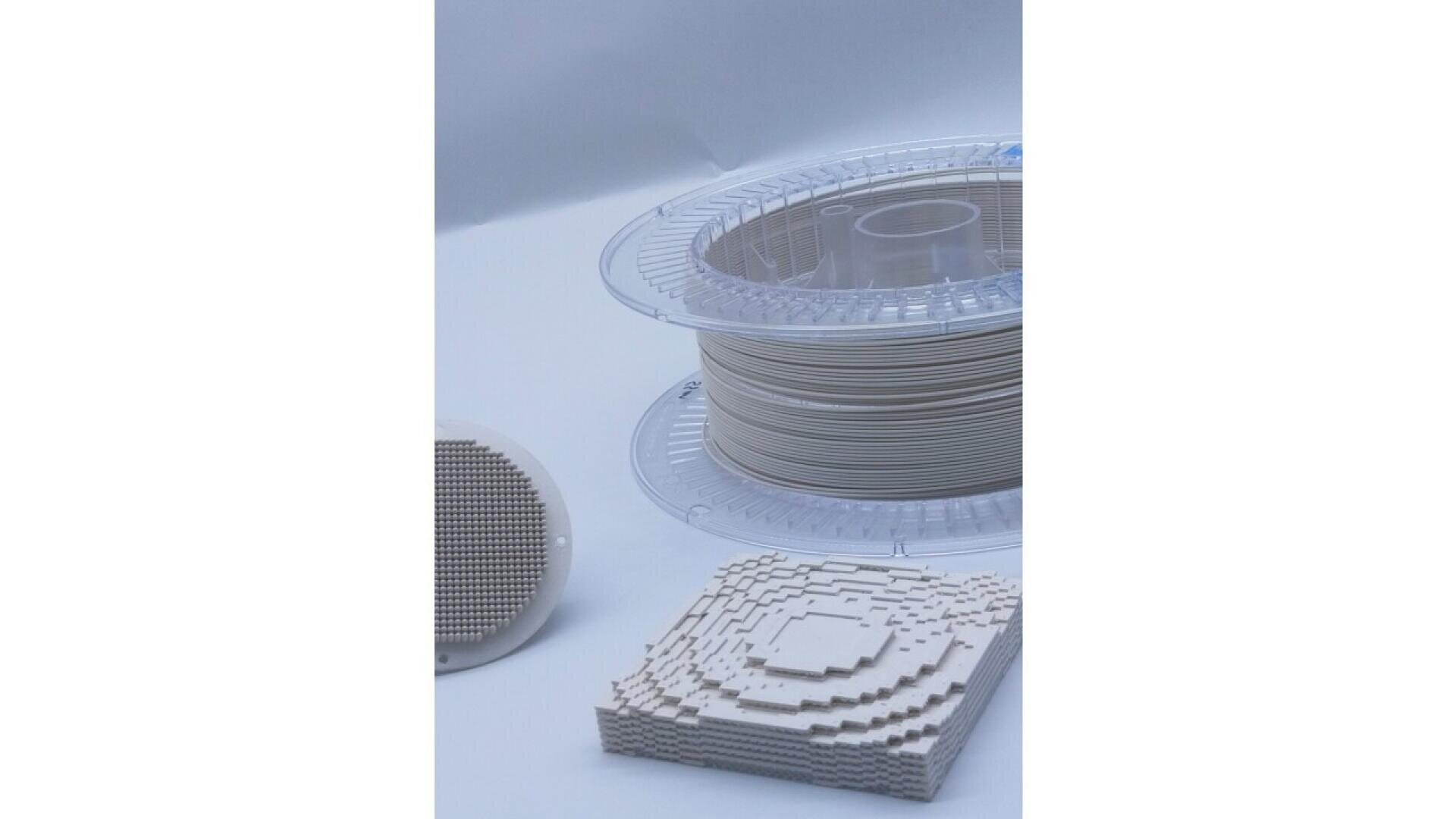 A spool of beige 3D printing filament sits beside two 3D-printed objects: a circular grid and a stacked, layered square structure, all displayed on a white surface.