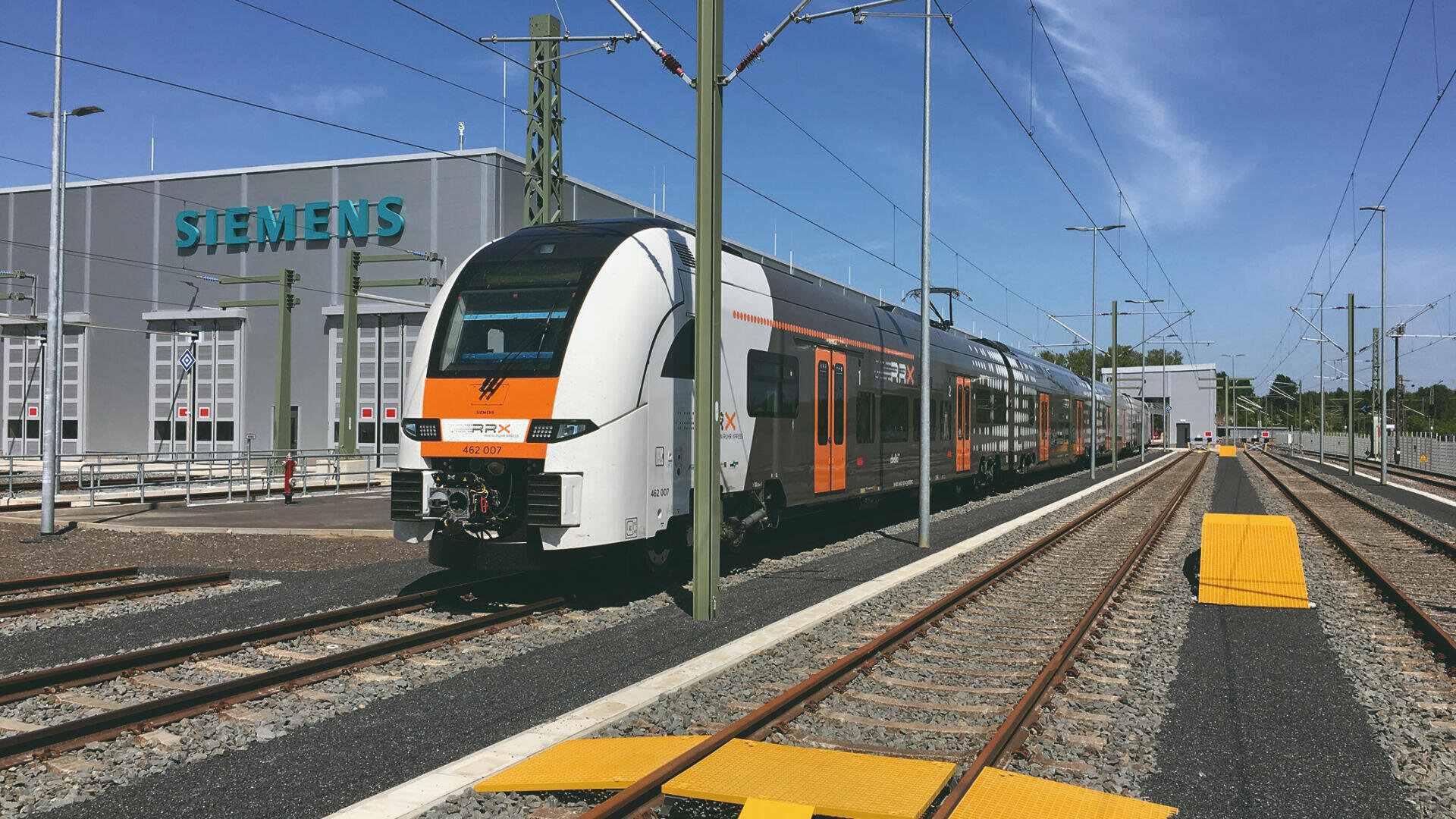 Ein moderner orange-weiß-schwarzer Personenzug fährt unter einem klaren blauen Himmel auf den Gleisen vor einem Siemens-Gebäude, umgeben von Stromleitungen und Infrastruktur.