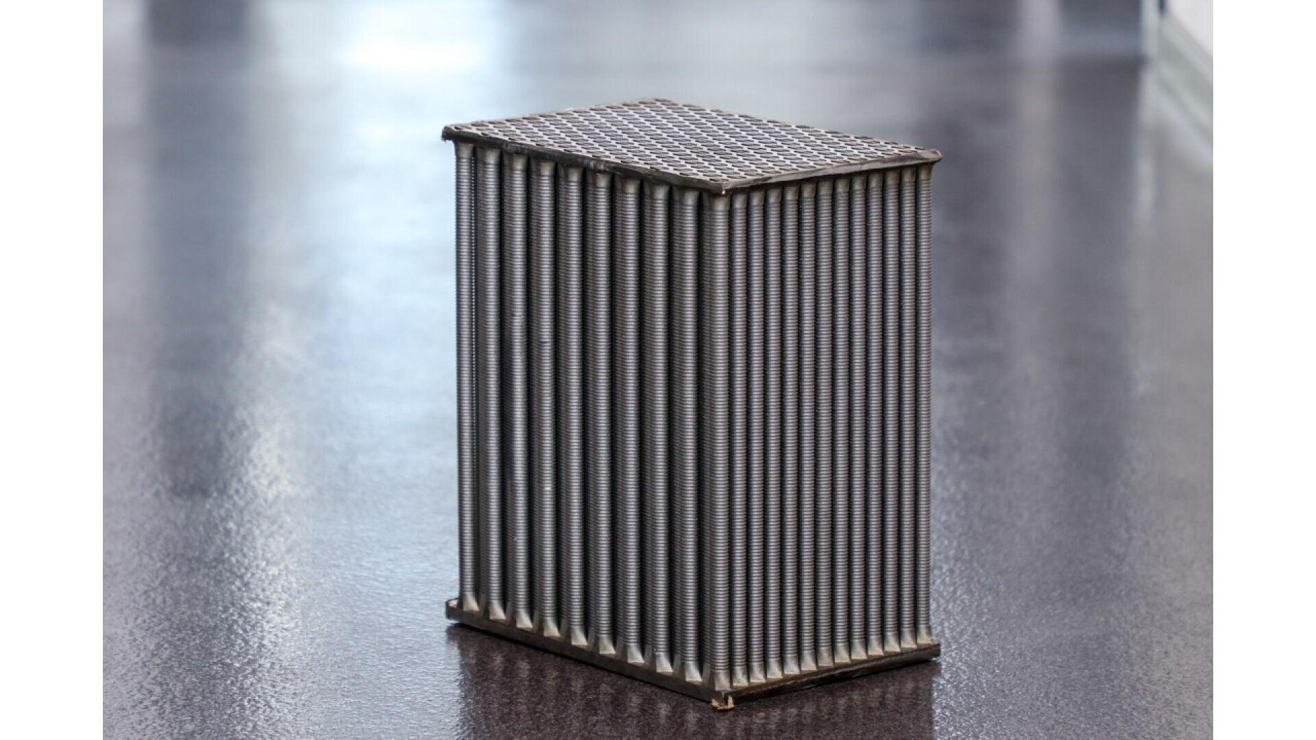 A rectangular block with a grid-patterned top and vertically aligned metallic rods on its sides, sitting on a smooth, reflective floor.