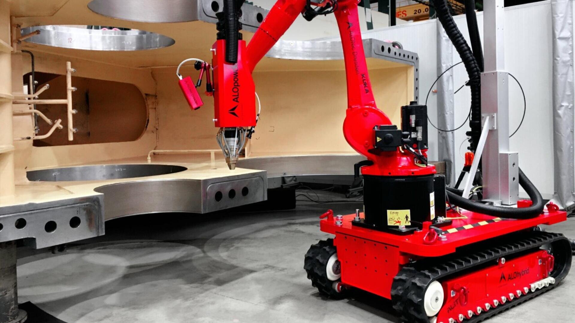 A red robotic arm mounted on a tracked base works on a large, partially assembled industrial structure in a factory setting. The robot appears to be engaged in a construction or manufacturing task.