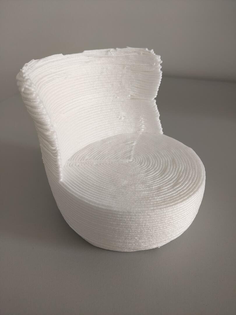 Ein kleiner, weißer 3D-gedruckter Stuhl mit abgerundeter Sitzfläche und geschwungener Rückenlehne, der durch den Druckprozess eine sichtbare Schichtstruktur aufweist, steht auf einer schlichten grauen Fläche.