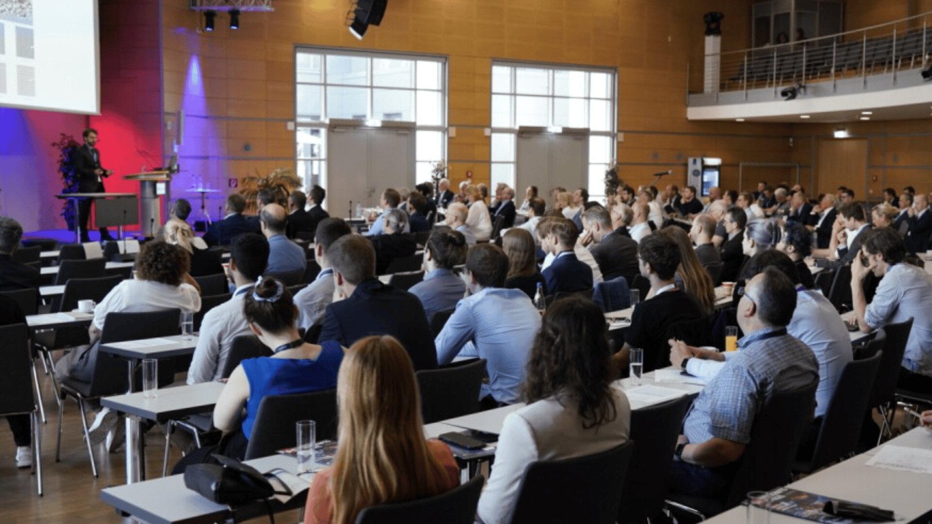 A large group of people sits at tables facing a stage during a conference or seminar in a modern, well-lit room. A speaker stands at the podium, presenting to the attentive audience.