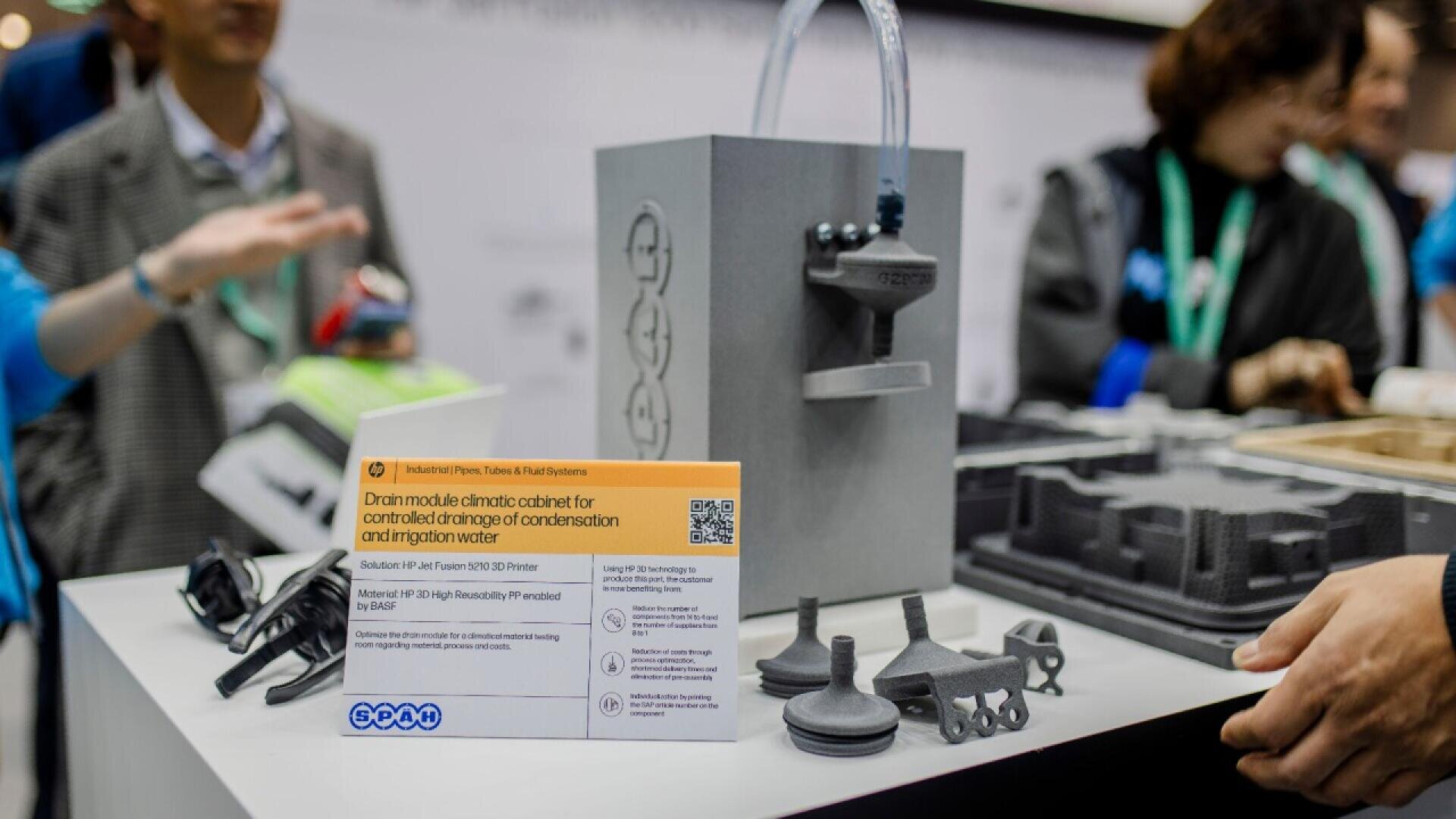 A display table shows a gray climate cabinet model with attached tubes, several 3D-printed parts, an informational sign, and blurred people interacting in the background at an exhibition.