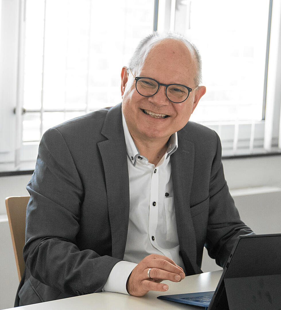 Ein lächelnder älterer Mann mit Brille und grauem Anzug sitzt mit einem Laptop an einem Schreibtisch in einem hellen Büro mit großen Fenstern hinter ihm.
