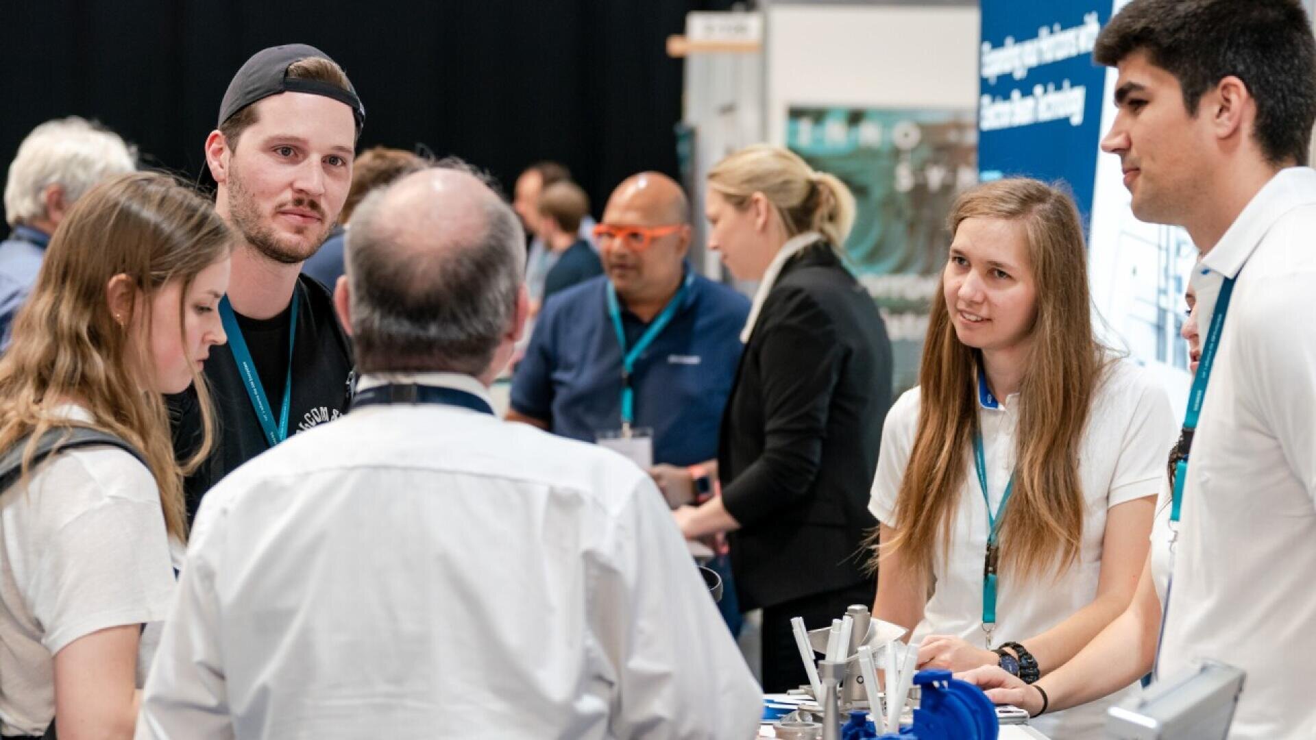 A group of young adults and an older man engage in conversation at a busy exhibition booth, with others networking and displays visible in the background.