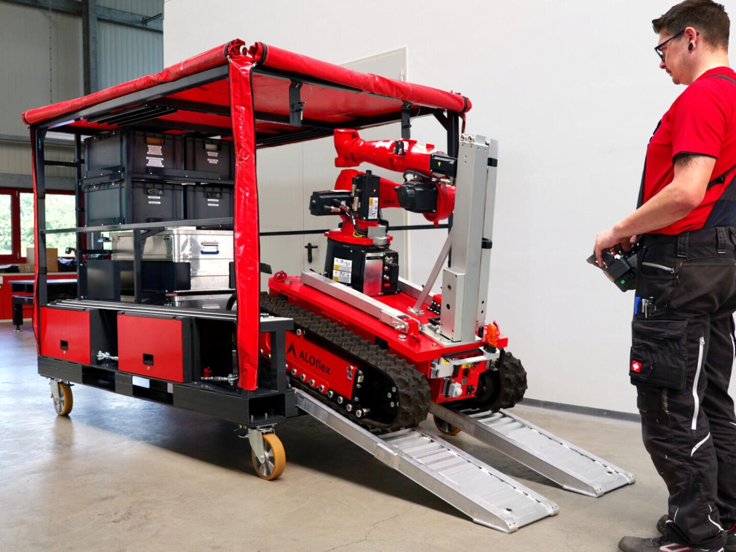 A person in a red shirt operates a tracked red robot with a mechanical arm as it drives down a metal ramp from a mobile storage cart inside a building.