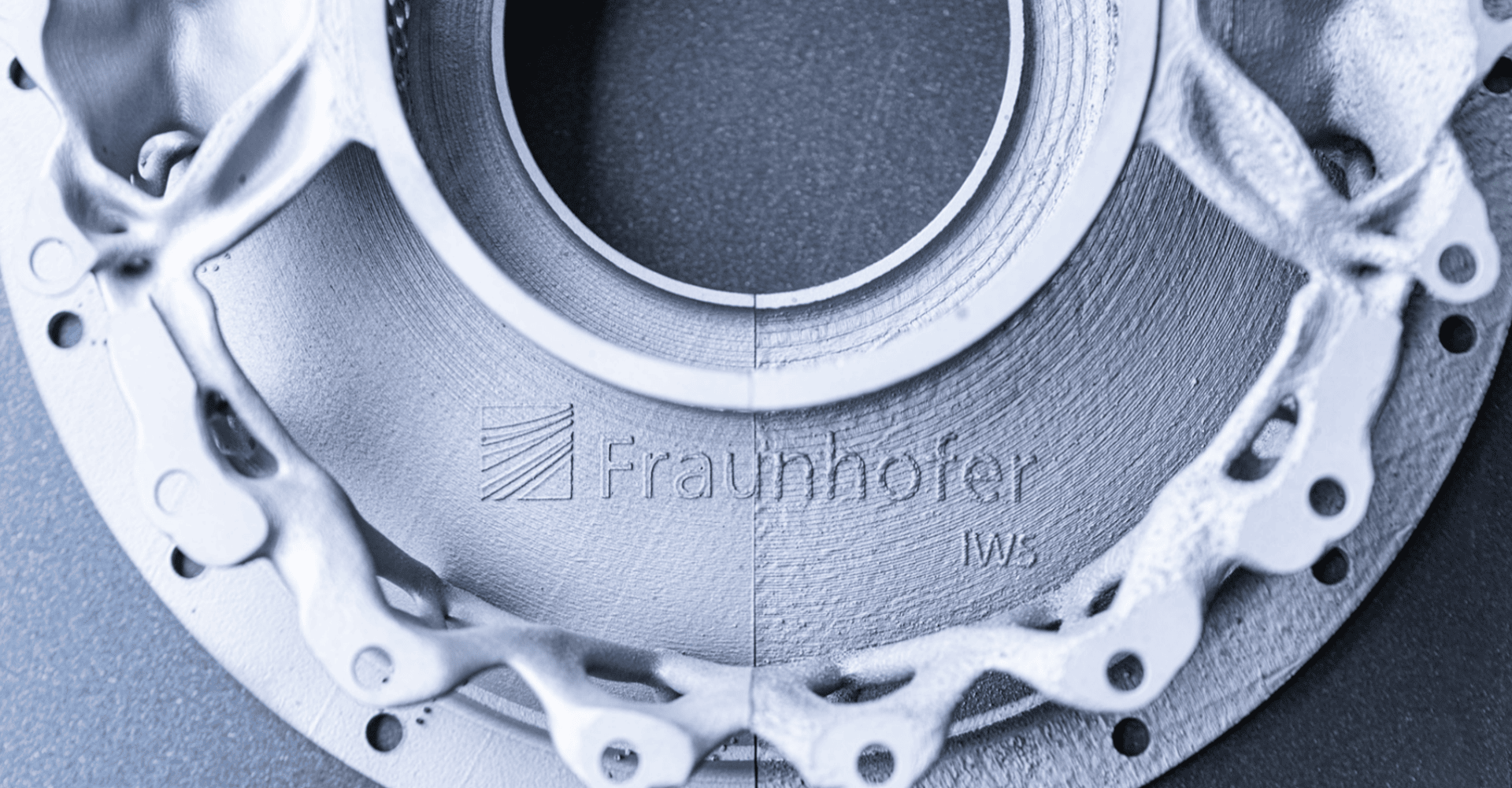 Nahaufnahme eines metallischen, kreisförmigen Maschinenteils mit komplexen Rippen und Löchern. Der Schriftzug „Fraunhofer IWS“ ist in die Oberfläche eingraviert. Das Bild hat einen blauen Farbton und zeigt feine Oberflächendetails.