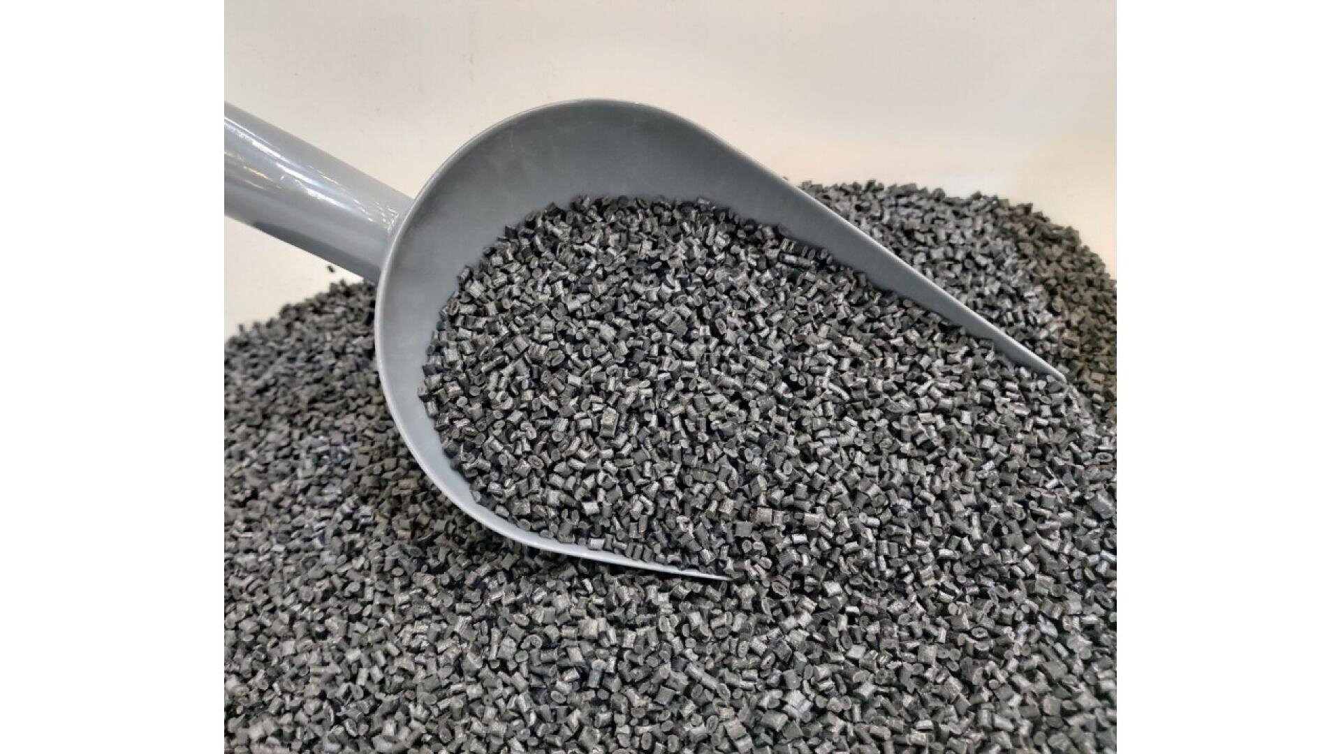A gray scoop filled with small, dark gray plastic pellets, with more pellets spread across the surface below.