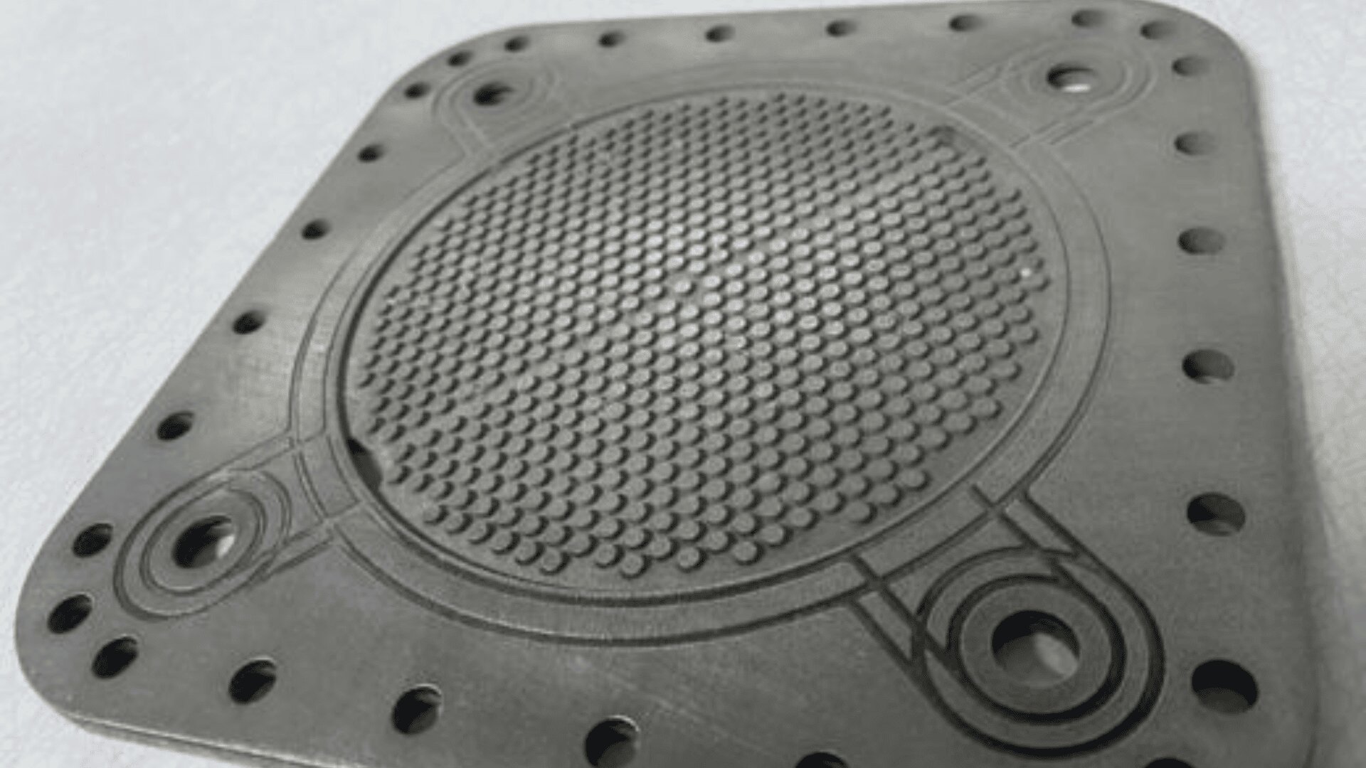 A close-up of a metallic, square-shaped plate with rounded corners, evenly spaced holes along its edges, and a textured circular pattern in the center. The surface also features several engraved lines and circular designs.