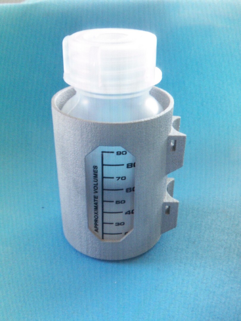 Eine kleine durchsichtige Plastikflasche mit weißem Deckel, gehalten in einem grauen Halter mit einer ungefähren Volumenskala von 30 bis 90 Milliliter, platziert auf einem blauen strukturierten Hintergrund.