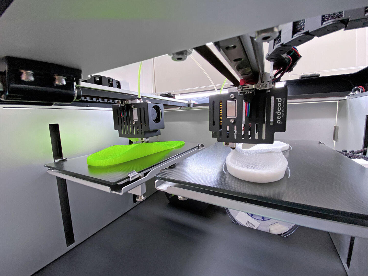 Eine Nahaufnahme eines 3D-Druckers im Betrieb mit zwei Druckköpfen, die Objekte produzieren; einer druckt ein grünes Objekt und der andere stellt auf benachbarten Plattformen ein weißes Objekt her.