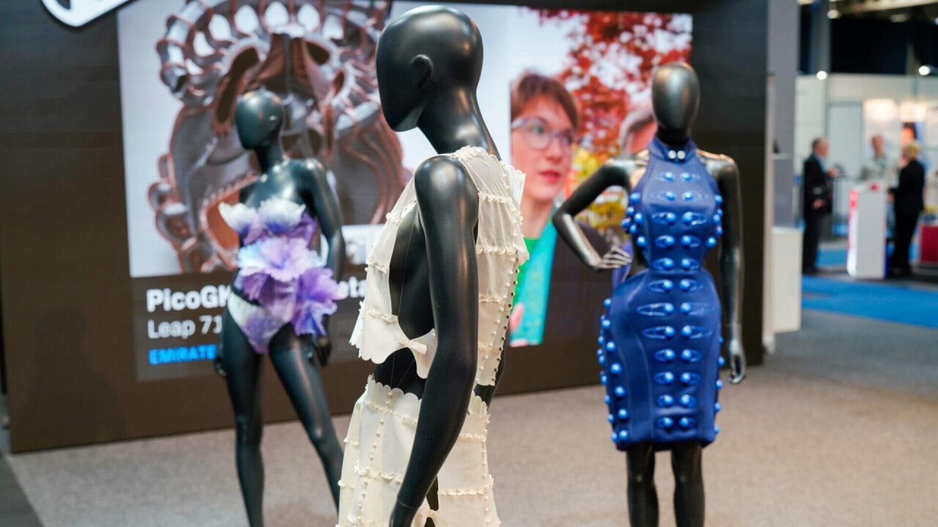 Three mannequins display futuristic, 3D-printed fashion designs at an exhibition. In the background, a large digital screen shows a womans face and abstract graphics, with booths and visitors visible in the distance.